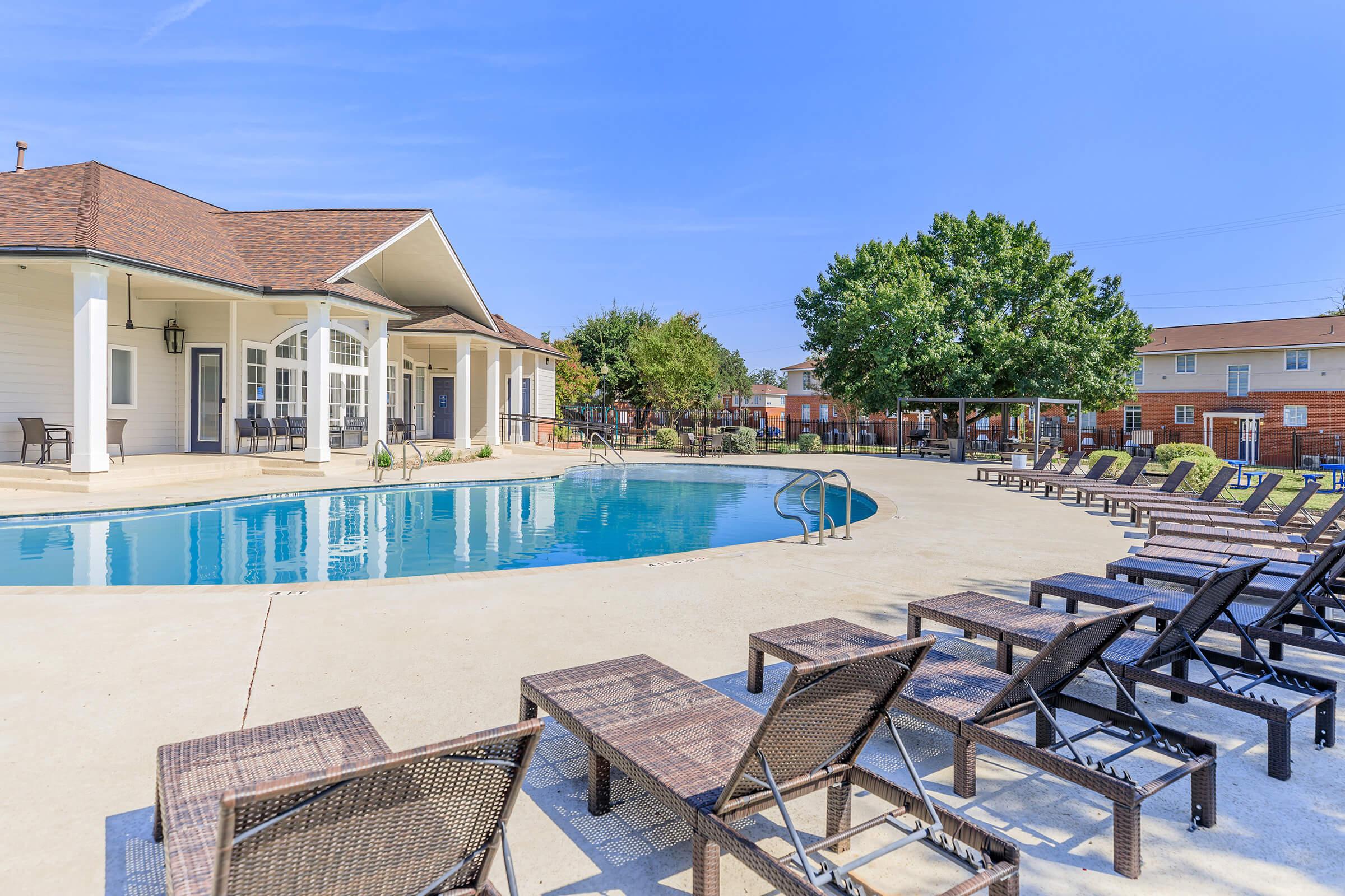 A serene pool area featuring a swimming pool surrounded by lounge chairs, with a fenced yard, green trees, and a residential building in the background under a clear blue sky. Perfect for relaxation and outdoor enjoyment.