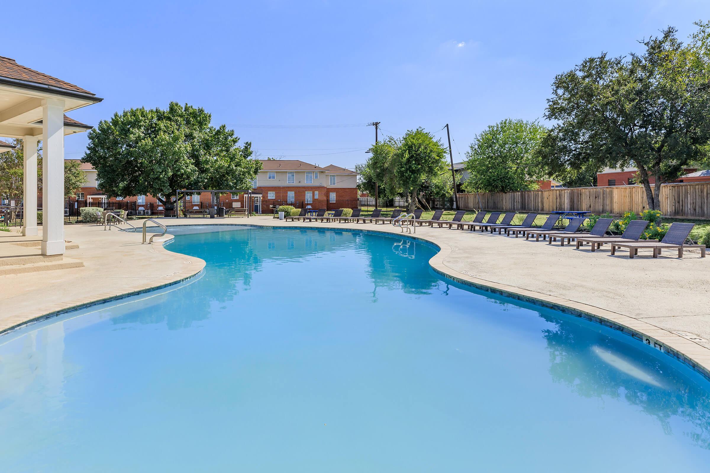 A clear blue swimming pool surrounded by lounge chairs and trees, set in a residential community. The pool area features a deck with umbrellas and a cozy atmosphere, perfect for relaxation. Bright sunlight reflects off the water, creating a serene outdoor environment.