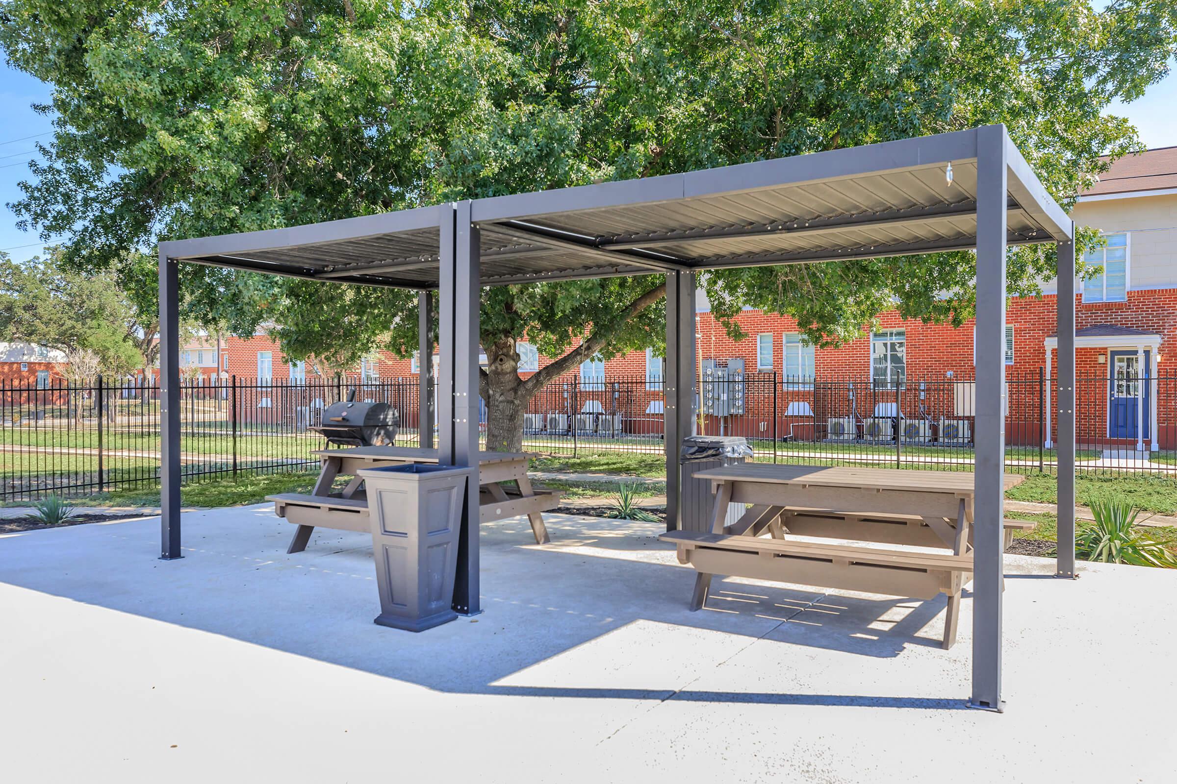 Outdoor picnic area featuring a covered shelter with two picnic tables and a grill. The setting includes green trees and buildings in the background, providing a welcoming space for gatherings or meals. The surface is concrete, and there is a fence around the area.