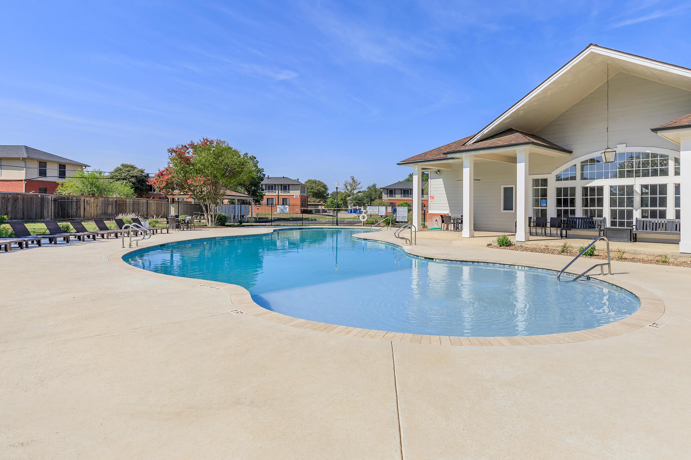 A modern swimming pool surrounded by a concrete patio, featuring lounge chairs on one side. The pool area is complemented by a spacious clubhouse with large windows and a gabled roof. Lush greenery and residential buildings are visible in the background under a clear blue sky.