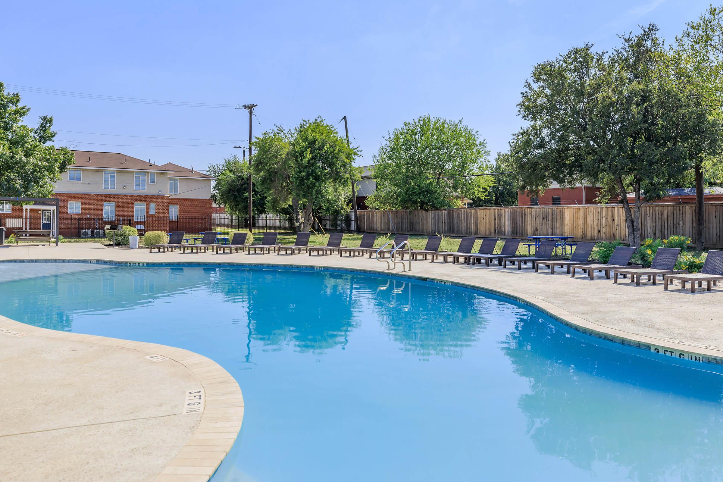 A tranquil outdoor swimming pool surrounded by lounge chairs, set against a clear blue sky. Lush trees provide shade near the pool area, while a wooden fence encloses the space. Nearby buildings are visible in the background, creating a serene atmosphere ideal for relaxation.