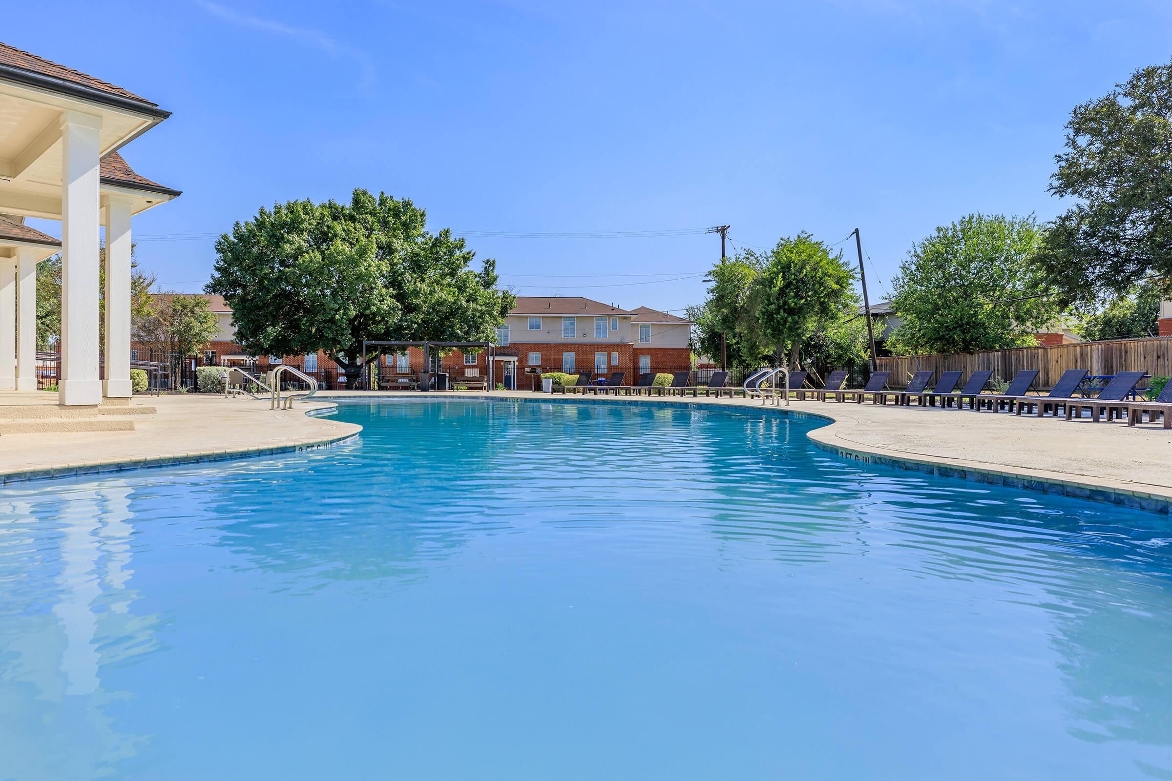 A large swimming pool with clear blue water, surrounded by patio furniture and trees. In the background, residential buildings are visible under a clear blue sky. The setting appears inviting and perfect for relaxation.
