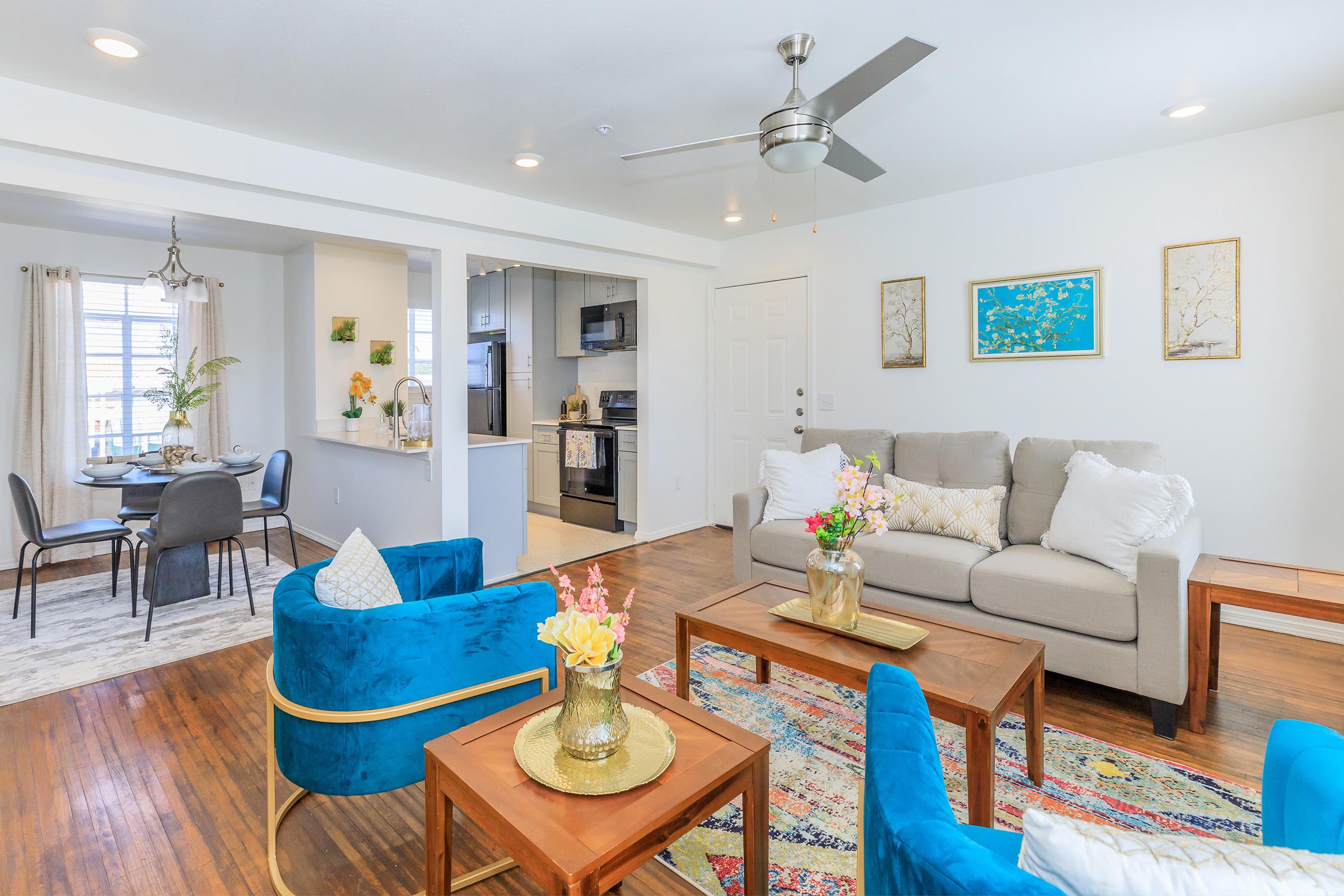 A bright and modern living room featuring a comfortable gray sofa, two blue armchairs, and wooden coffee tables with decorative vases of flowers. In the background, there’s a dining area with a round table and four chairs, and a stylish kitchen visible. The walls are adorned with framed artwork.