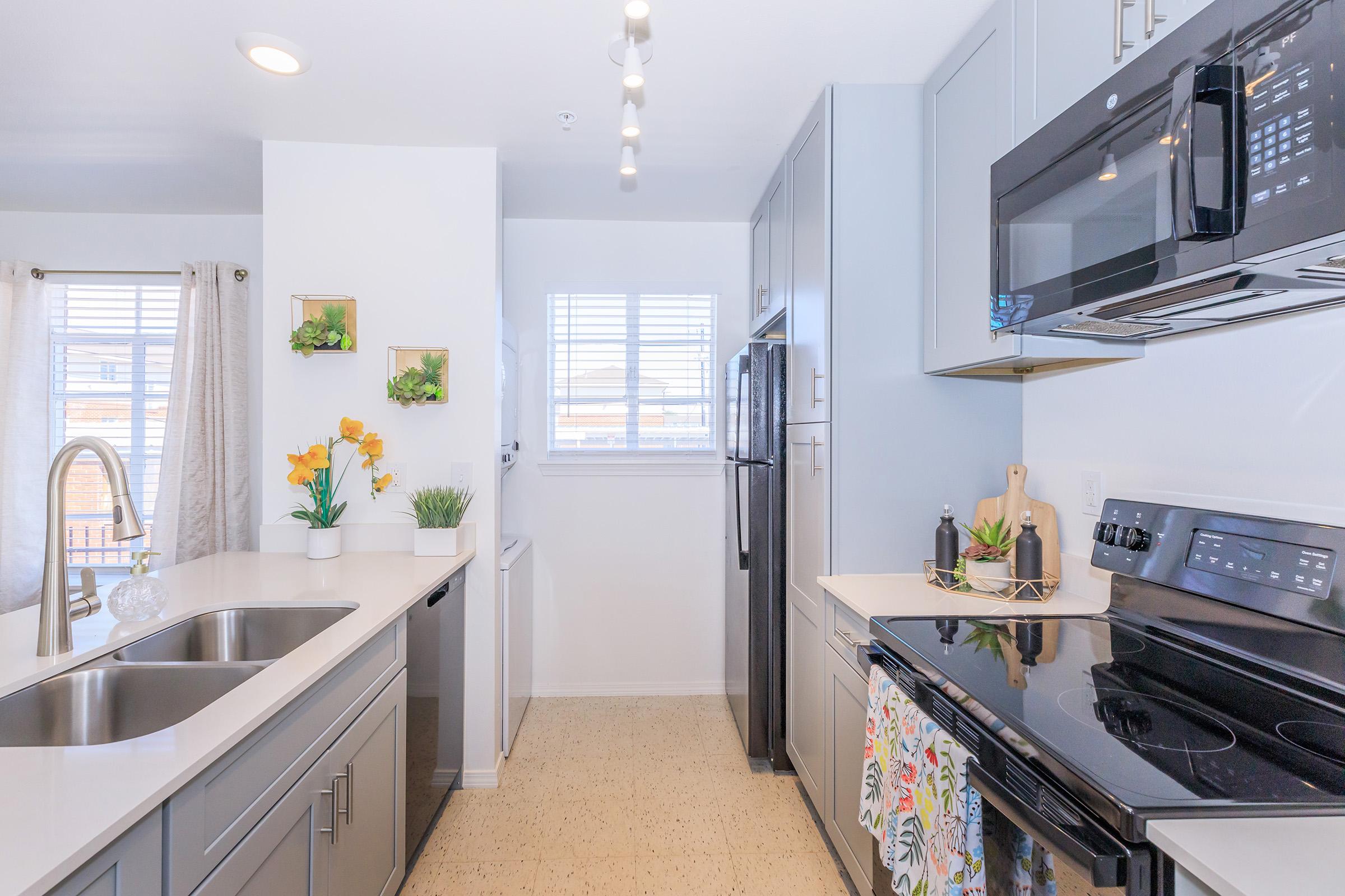 Modern kitchen featuring gray cabinets, stainless steel appliances, and a white countertop. A double sink is positioned under a window, with decorative plants and yellow flowers on display. The space is well-lit and organized, highlighting a microwave, oven, and refrigerator.