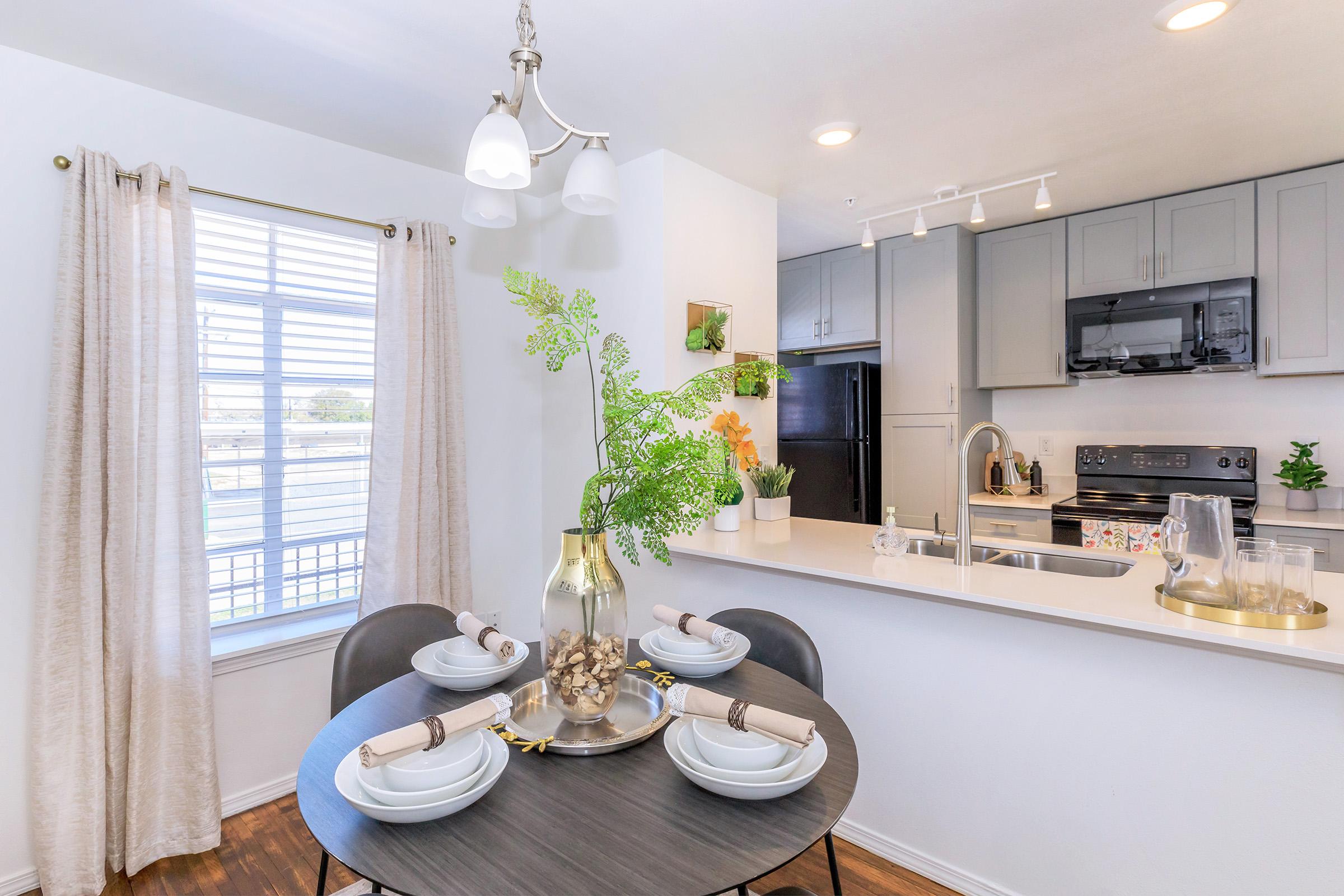 A modern kitchen and dining area featuring a round table set with four white plates and silver utensils. A stylish vase with greenery sits at the center. The kitchen has gray cabinets, a black stove, and an open layout with a window letting in natural light. Neutral curtains frame the window, adding a cozy touch.