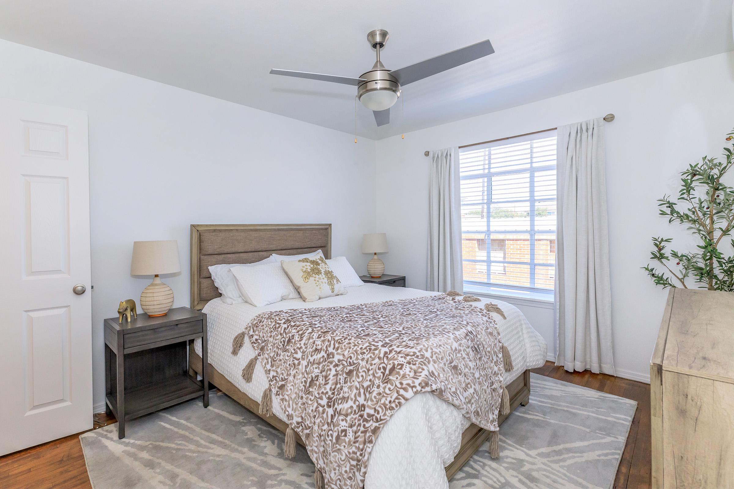A bright and cozy bedroom featuring a wooden bed with a light blanket, a bedside table with a lamp, and a window dressed with sheer curtains. The walls are painted white, and there's a decorative plant in the corner, enhancing the serene ambiance of the space.