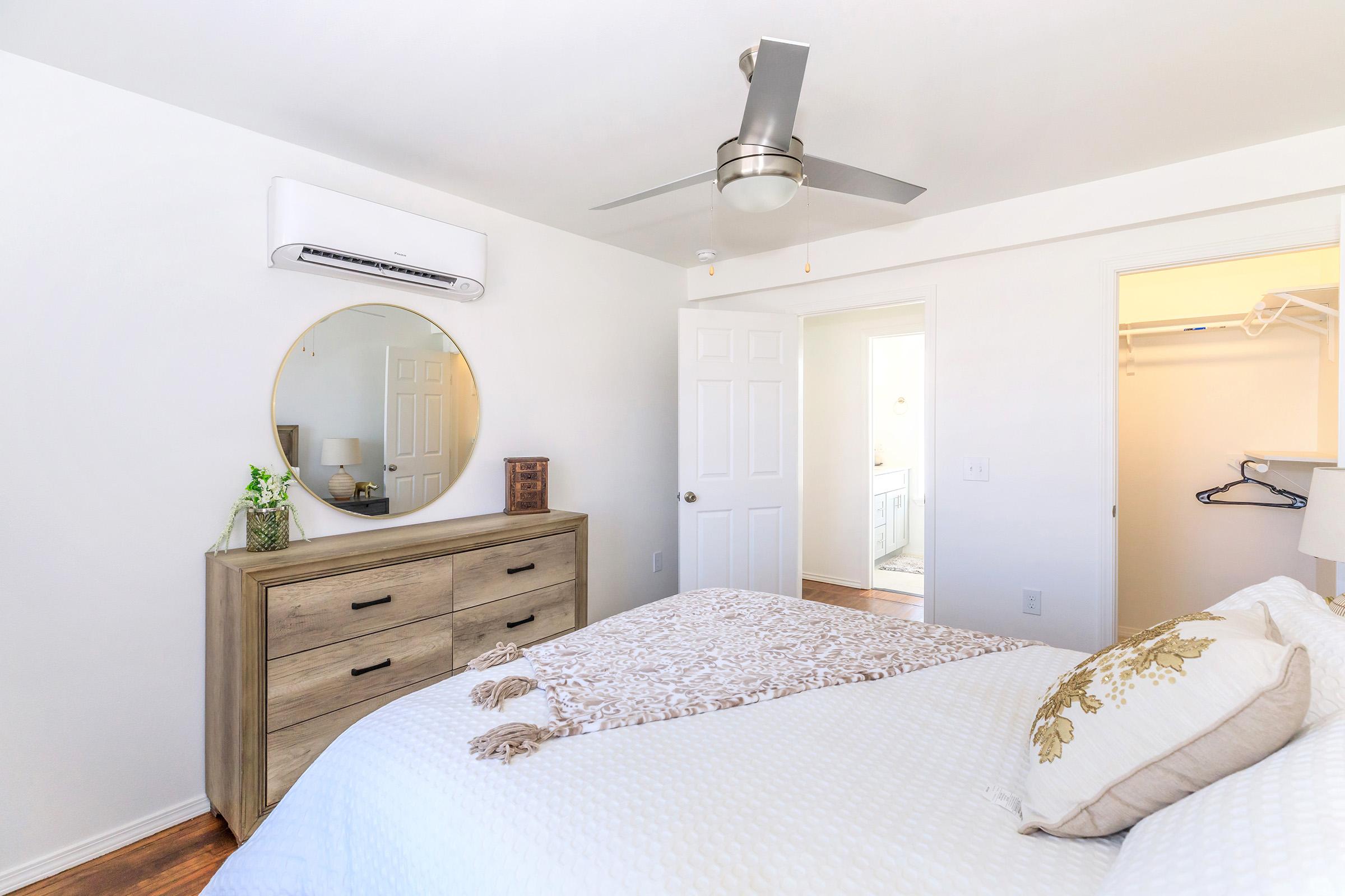 A modern bedroom featuring a large round mirror above a wooden dresser, a ceiling fan, and a neatly made bed with decorative pillows. A doorway leads to a closet and another room, and there is a small plant on the dresser, enhancing the clean and airy atmosphere.