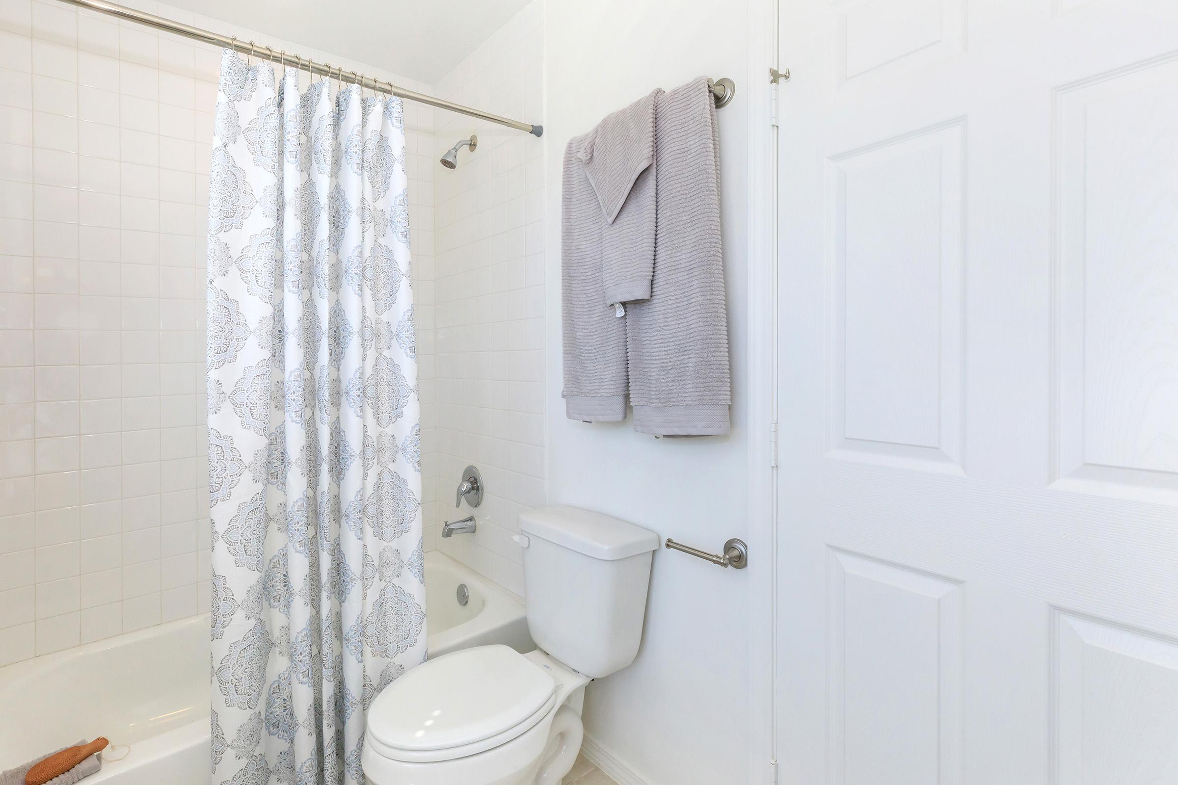 A clean bathroom featuring a white bathtub with a decorative shower curtain, a toilet, and grey towels hanging on a rack. The walls are light-colored, creating a bright and airy atmosphere. A white door is visible, adding to the minimalist design.