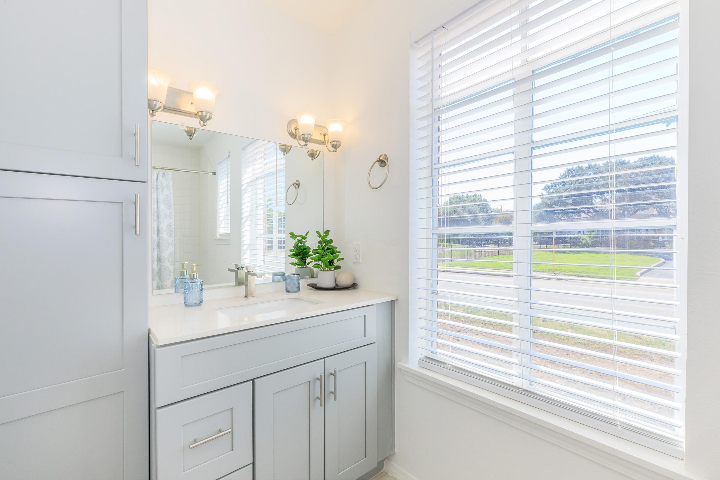 A modern bathroom featuring light gray cabinets, a white countertop, and a large mirror. Two light fixtures are mounted above the mirror. A small potted plant sits on the counter next to two glass containers. The window has white blinds, with a view of greenery outside.