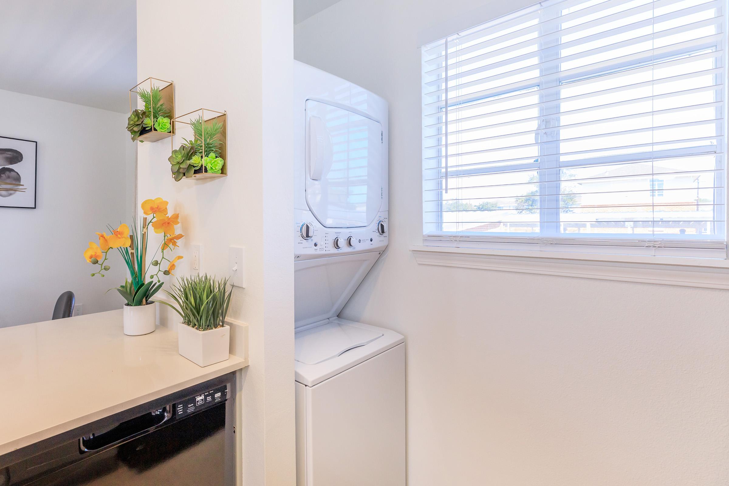 A bright laundry area featuring a stacked washer and dryer next to a window with blinds, adorned with potted plants and orange flowers on a countertop. The walls are painted in light colors, creating an airy and inviting atmosphere.
