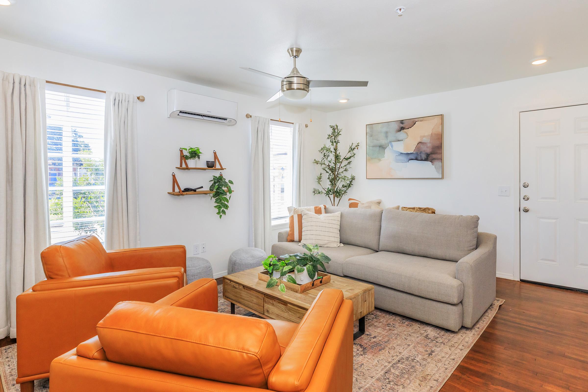 A modern living room featuring a light gray sectional sofa, two orange accent chairs, a wooden coffee table with plants on it, and a wall-mounted shelf with greenery. There are white curtains on windows, a ceiling fan, and a door leading outside. The decor is stylish and inviting.