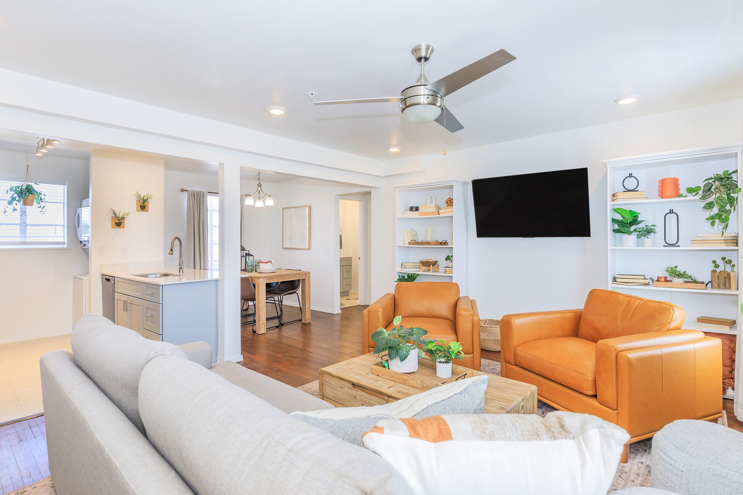 A modern living room featuring a light gray sectional sofa, two orange armchairs, and a wooden coffee table with plants. The space has a ceiling fan, wall-mounted TV, and built-in shelves decorated with greenery and accessories. A dining area is visible in the background with a table and chairs.