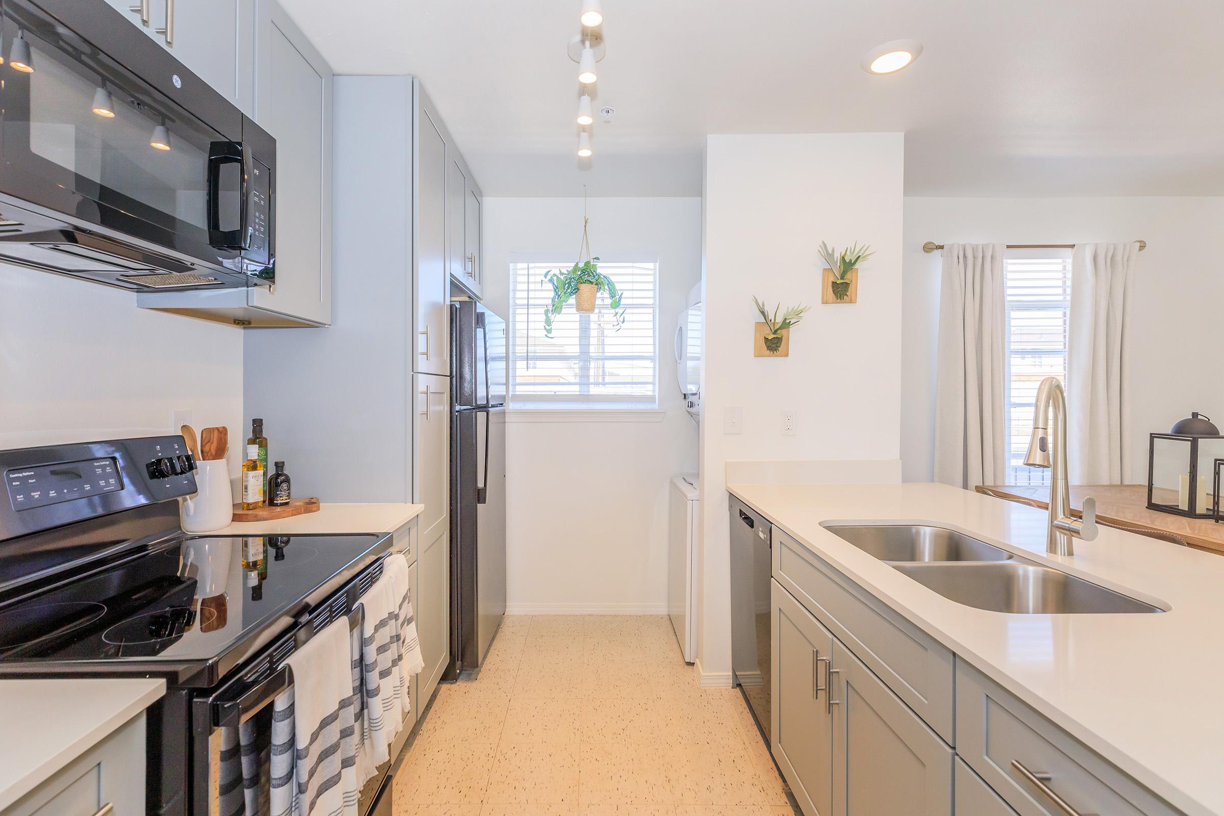 A modern kitchen featuring gray cabinets, stainless steel appliances, and a white countertop. The setup includes a microwave, oven, and refrigerator, with a window providing natural light. Decorative plants are mounted on the wall, enhancing the aesthetic. The space is clean and well-organized.