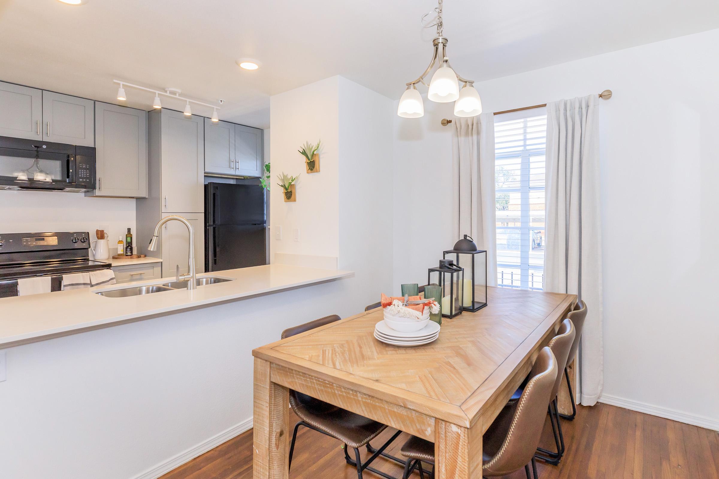 A modern kitchen and dining area featuring a wooden dining table set with plates and a lantern, surrounded by brown dining chairs. The kitchen has gray cabinets, stainless steel appliances, and a large window with white curtains allowing natural light in.