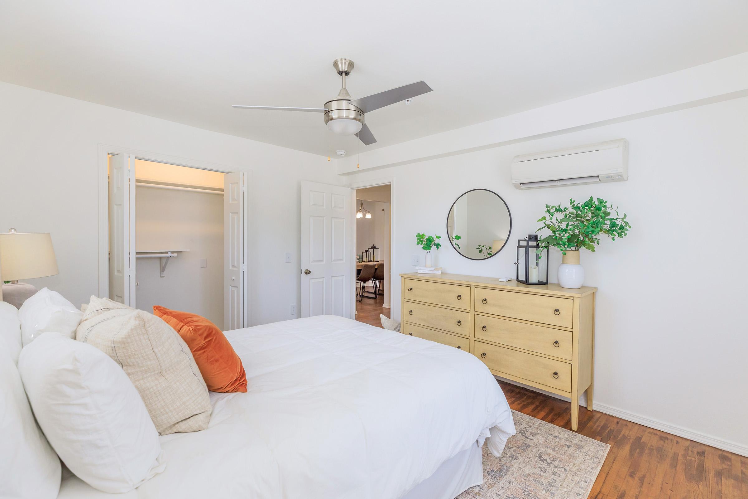 Bright and airy bedroom featuring a white bed adorned with orange and neutral pillows, a wooden dresser, a round mirror, and a ceiling fan. A closet door is visible in the background, along with indoor plants adding a fresh touch to the decor. Natural light enhances the inviting atmosphere.