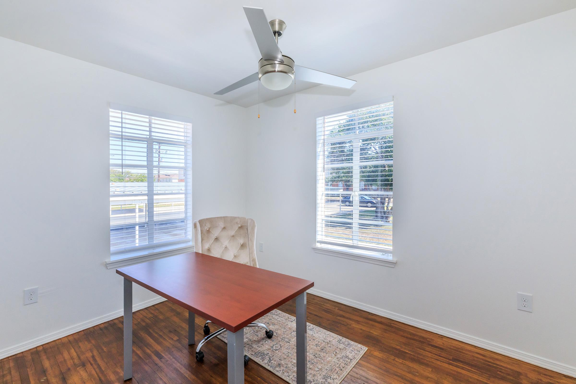 A bright and minimalistic office space featuring a wooden desk with a modern design, light-colored walls, two window panes allowing natural light, and a ceiling fan. The room has a comfortable chair and a small decorative rug on a wooden floor, creating a welcoming atmosphere for work or study.