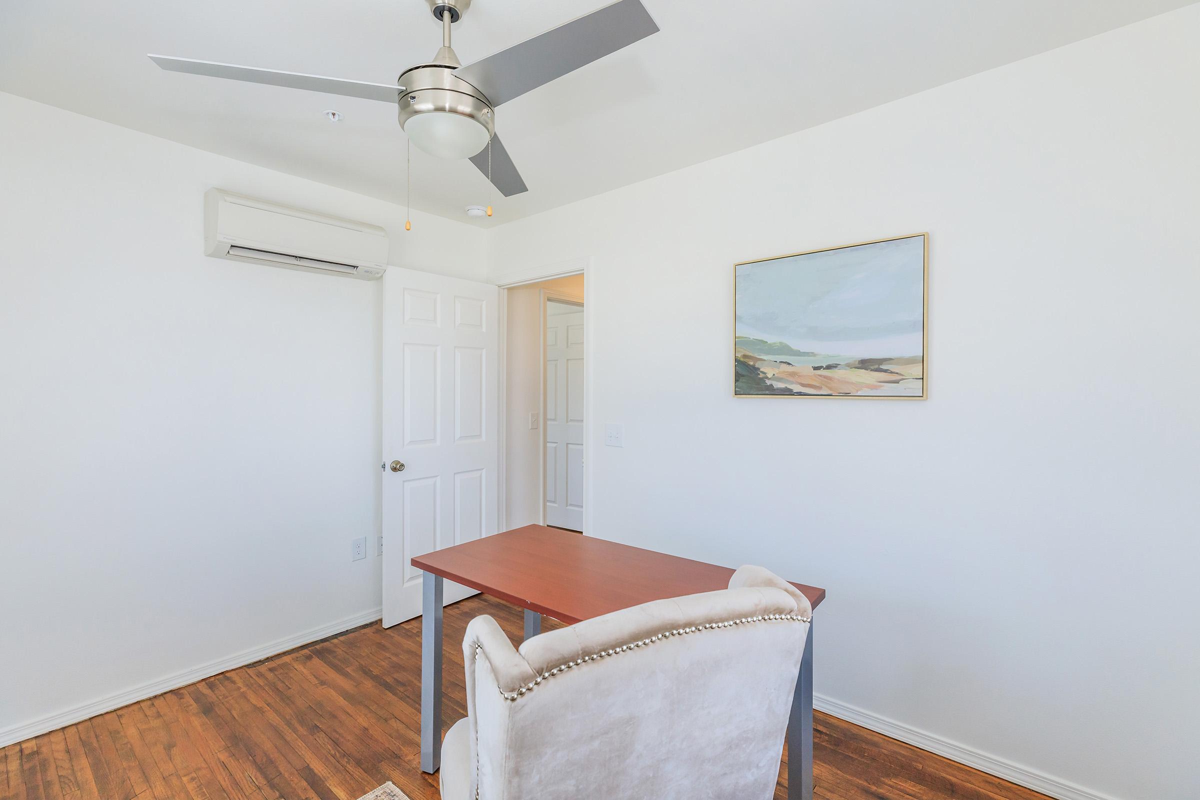 A bright and minimalistic room featuring a wooden desk with an upholstered chair, a ceiling fan, and a wall-mounted air conditioner. A painting of a landscape hangs on the wall, and a door leads to another room in the background. The clean, white walls and hardwood floor create a spacious feel.