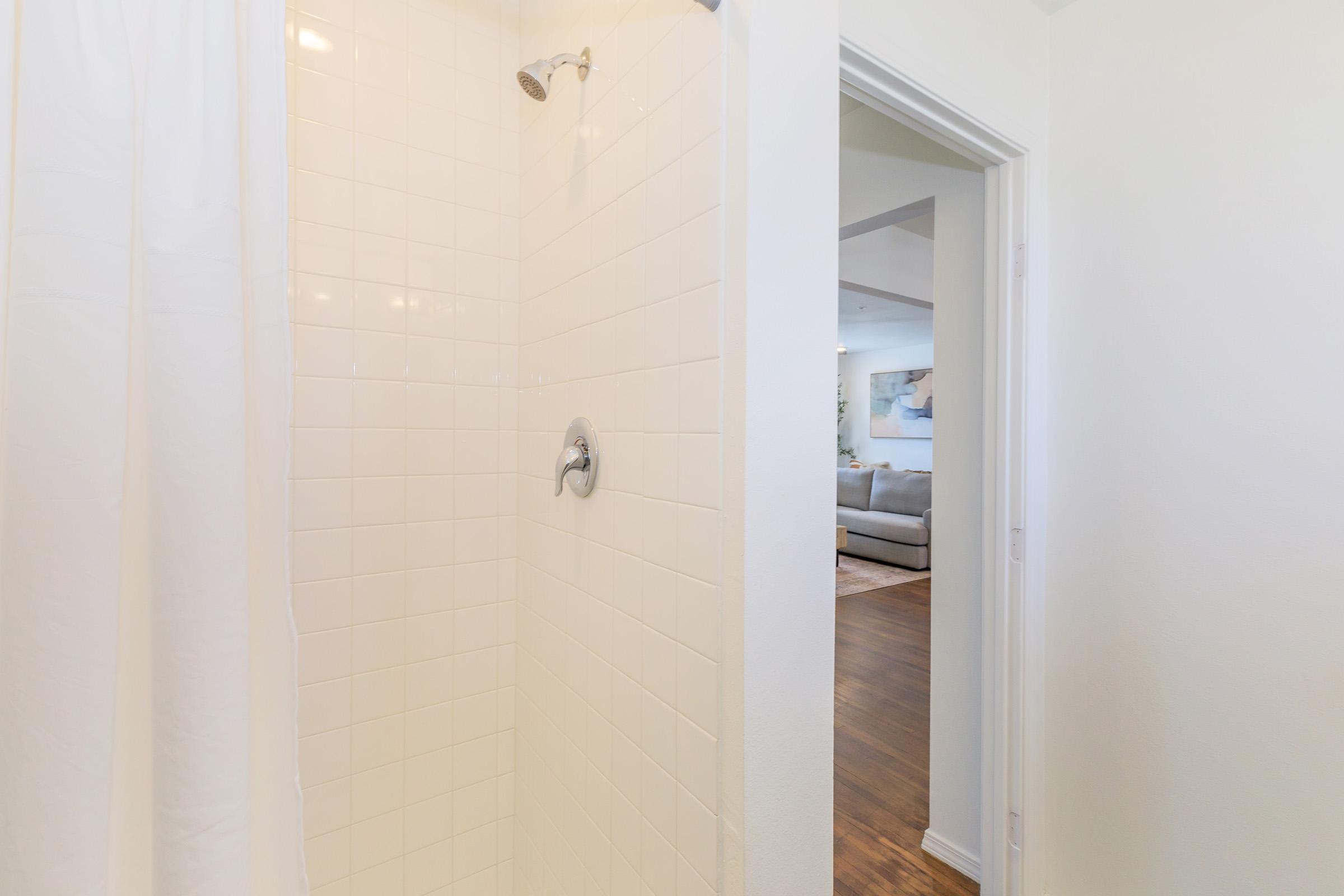 A clean, white tiled shower with a silver showerhead, partially obscured by a white shower curtain. In the background, there's an open doorway leading to a bright living area with a gray sofa and light-colored decor.