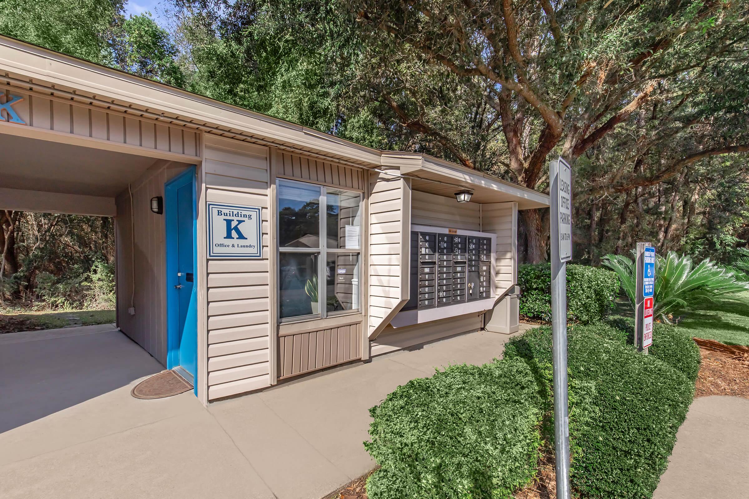 A low-rise building labeled "Building K" featuring a blue door, with a row of mailboxes on the side. The entrance is surrounded by greenery, including bushes and trees, creating a natural setting. A sign stands nearby, likely indicating directions or information related to the building.