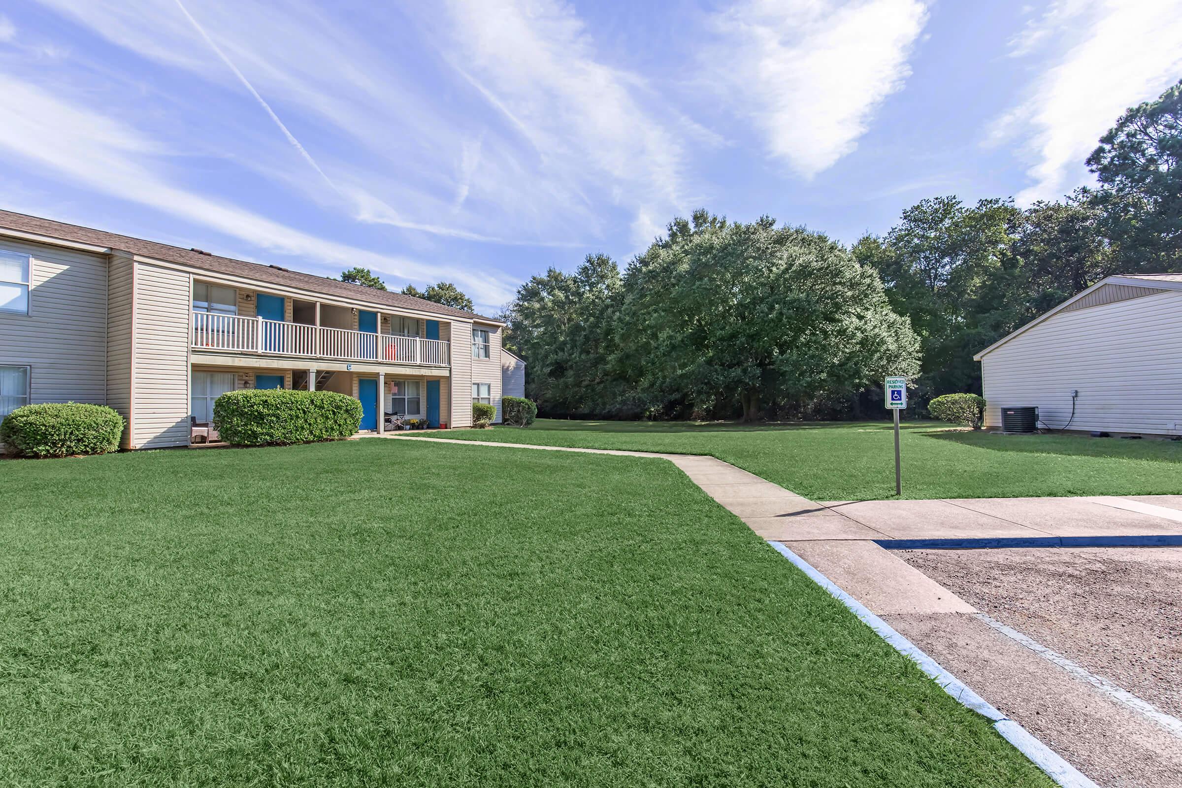 A view of a landscaped apartment complex with green grass and a pathway. The two-story buildings have balconies, surrounded by trees and shrubbery. A parking area is visible on the right side of the image, indicating a residential setting with well-maintained outdoor space.