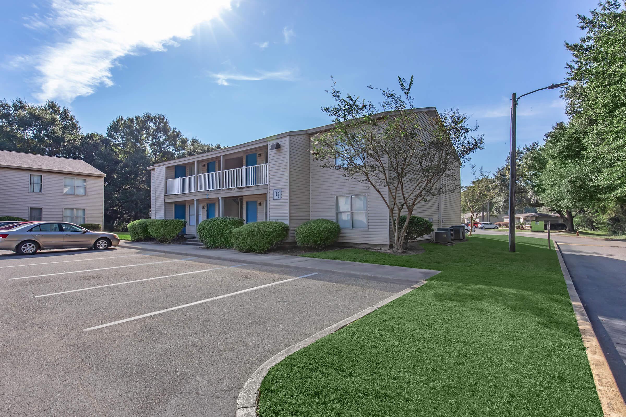 A two-story apartment building with light-colored siding, surrounded by greenery and trees. There are several parking spaces in front, and a clear blue sky overhead. A light post is visible beside the building, adding to the residential atmosphere.