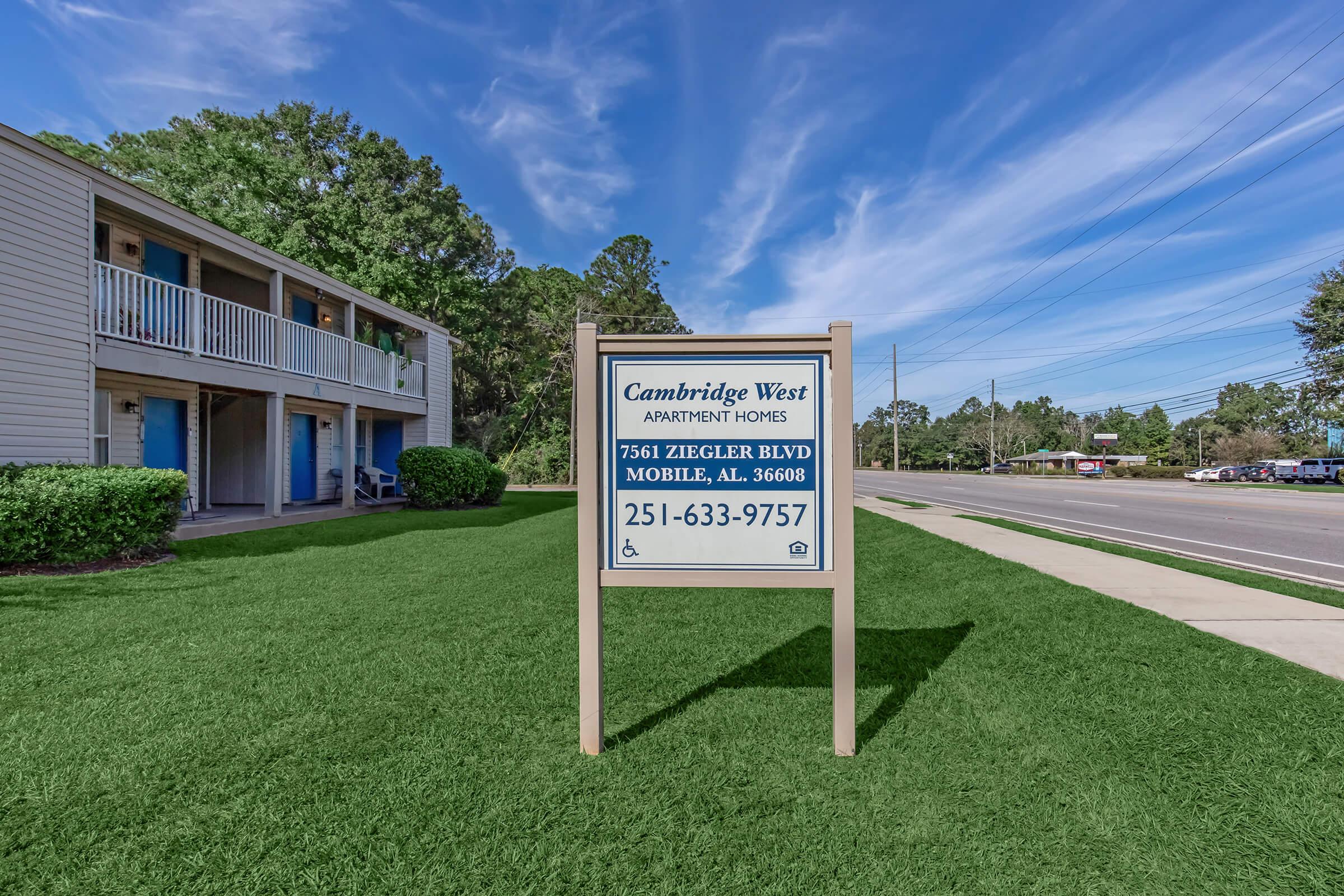 Sign for Cambridge West Apartment Homes located at 7561 Zeigler Blvd, Mobile, AL, with contact number 251-633-9757. The sign is situated in front of a well-maintained grassy area and a residential building under a clear blue sky.