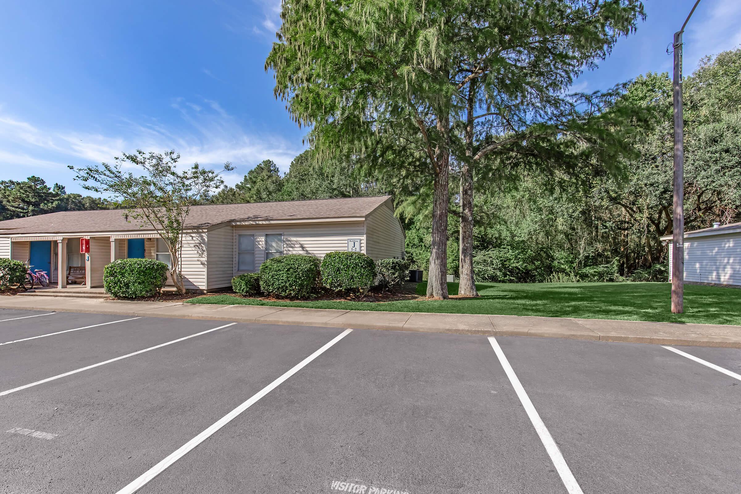A landscaped area featuring a single-story building with a porch and several bicycles parked in front. Trees and shrubs surround the building, and a grassy area is nearby. The parking lot has several empty spaces visible. The sky is clear with some clouds.