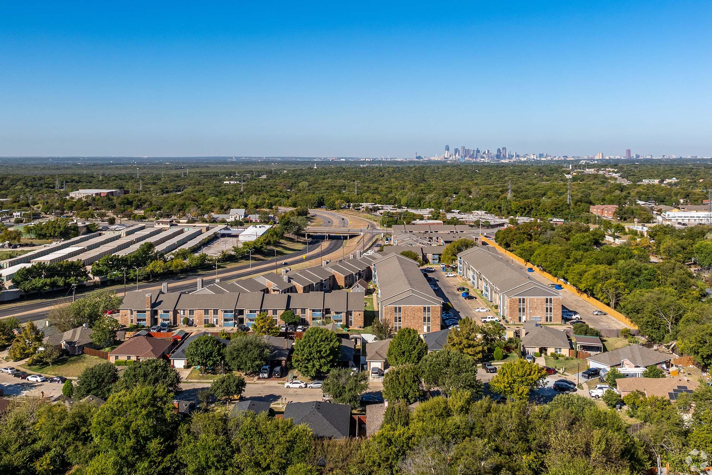 Aerial view of a suburban area featuring residential buildings, roads, and greenery, with a distant skyline of a city under a clear blue sky. The foreground shows multiple apartment complexes, while the background reveals urban structures on the horizon.