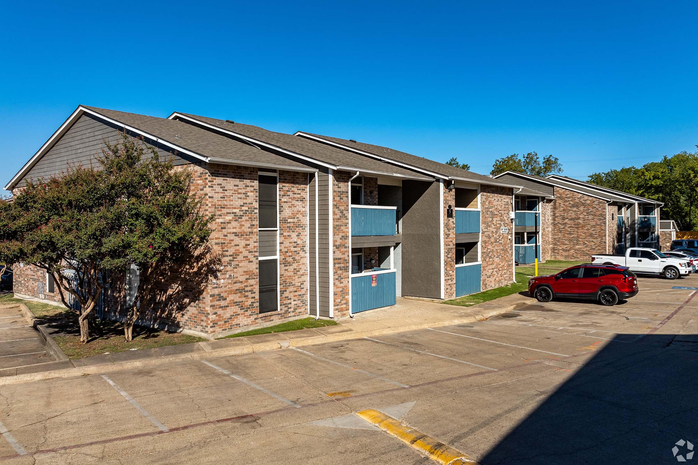 A multi-story apartment complex with a brick and blue exterior. The building features multiple units, balconies, and windows. The parking lot in front has a few vehicles, including a red car and a white vehicle. The scene is set against a clear blue sky.