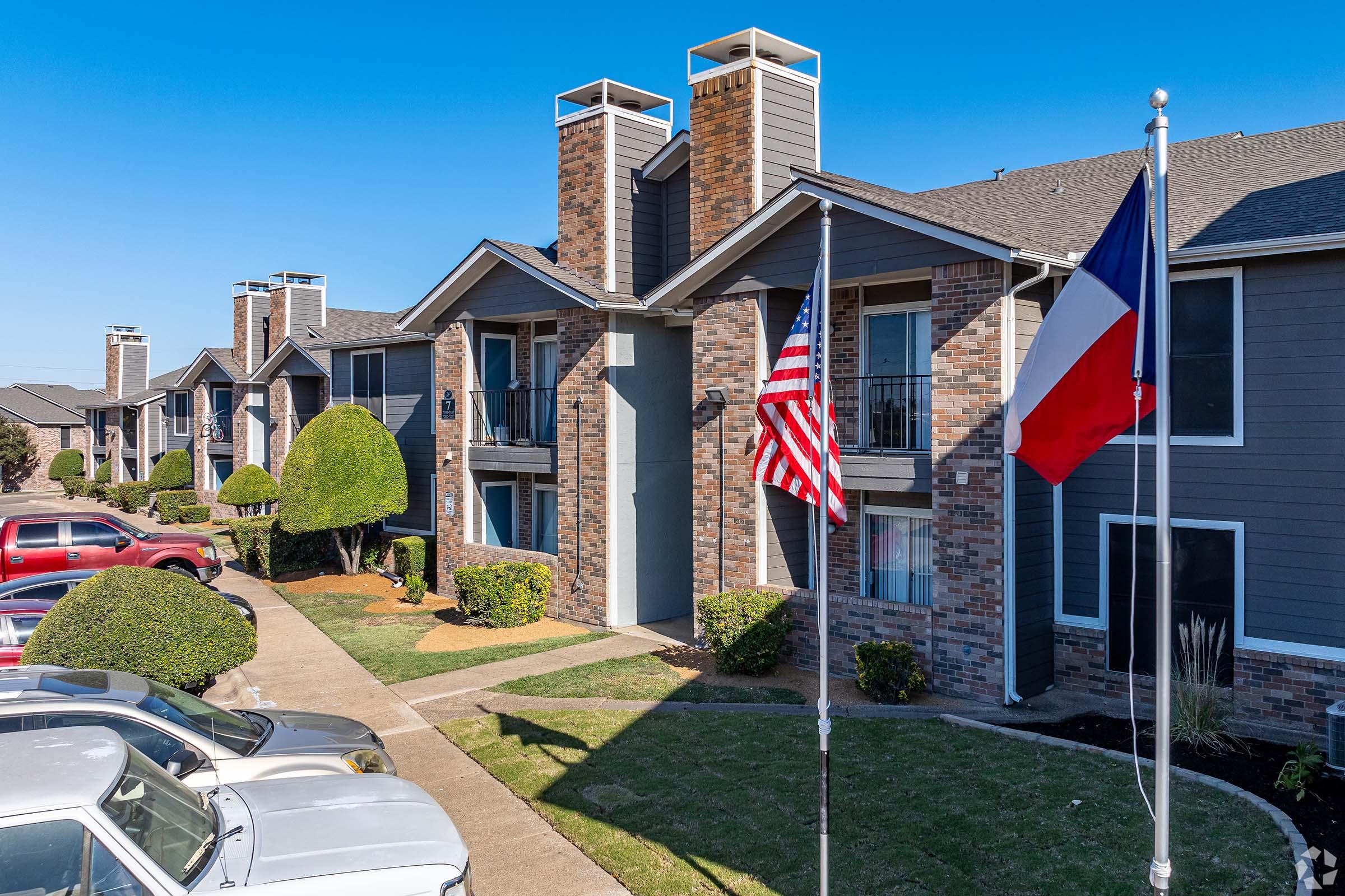 A view of a residential apartment complex featuring two buildings with brick facades and a well-manicured lawn. Two flags are prominently displayed: one American flag and one Texas state flag. Several parked cars are visible in the foreground, under a clear blue sky.