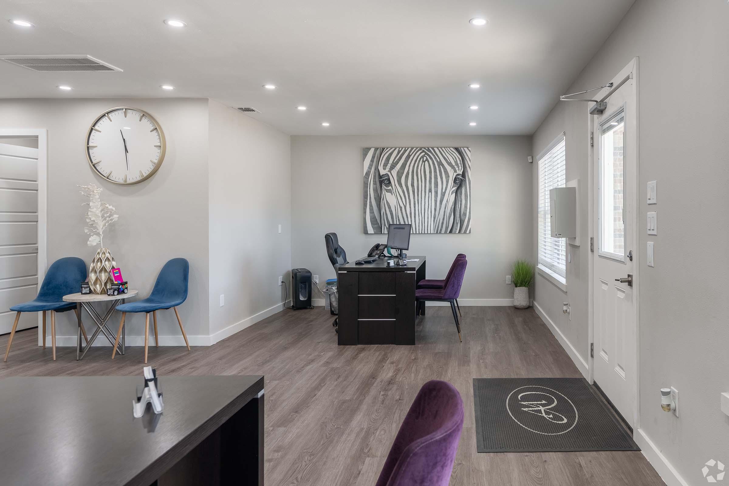 A modern office space featuring a desk and chair, a large clock on the wall, blue and purple upholstered chairs, and a decorative zebra print artwork. Natural light streams in through a window, highlighting the sleek hardwood floors and minimalist decor.