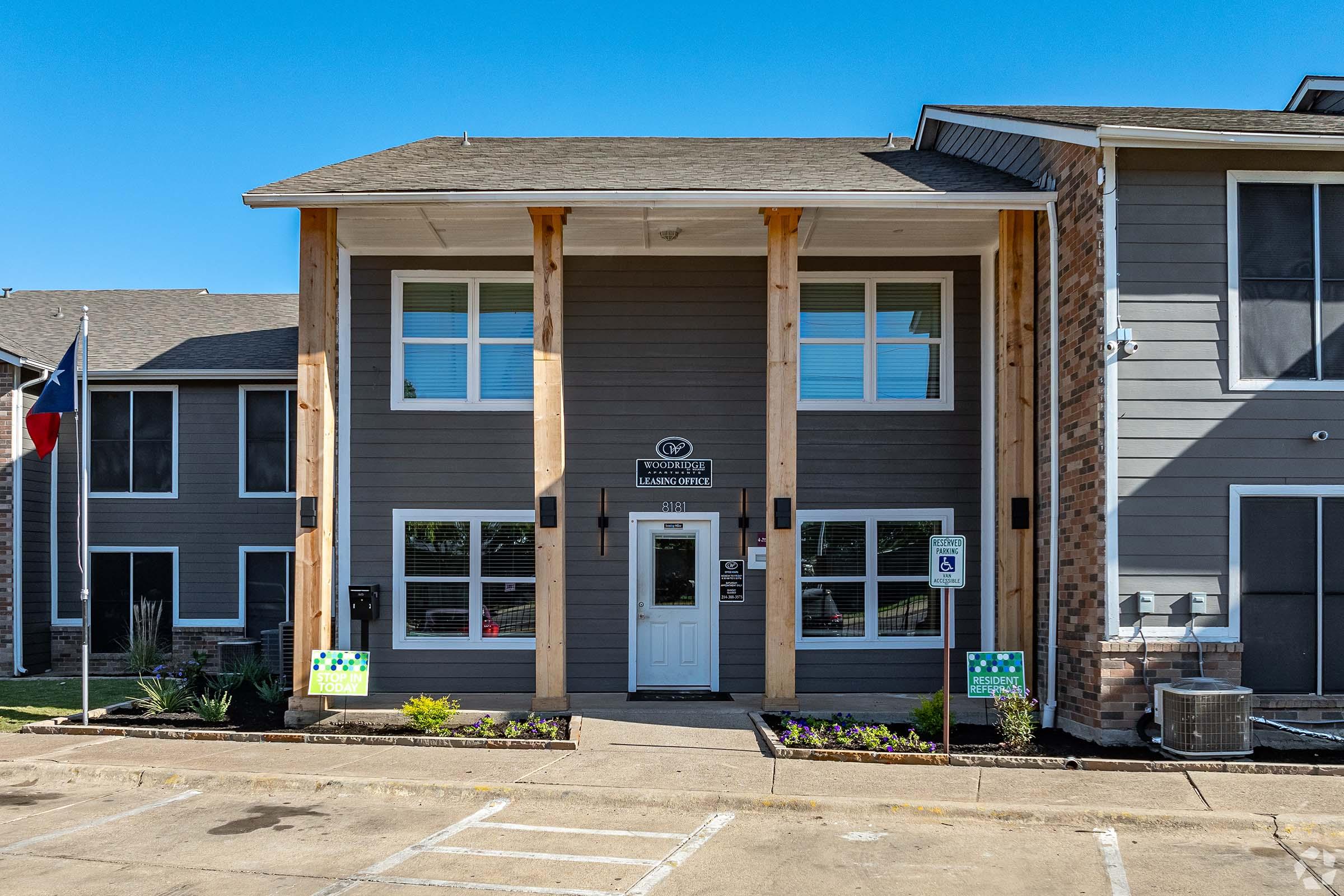 A modern apartment complex entrance featuring a two-story building with large windows, wooden pillars, and a front door. The exterior is a combination of gray siding and brick. There are parking spaces in front, with decorative landscaping including plants. A Texas flag is visible on the left.