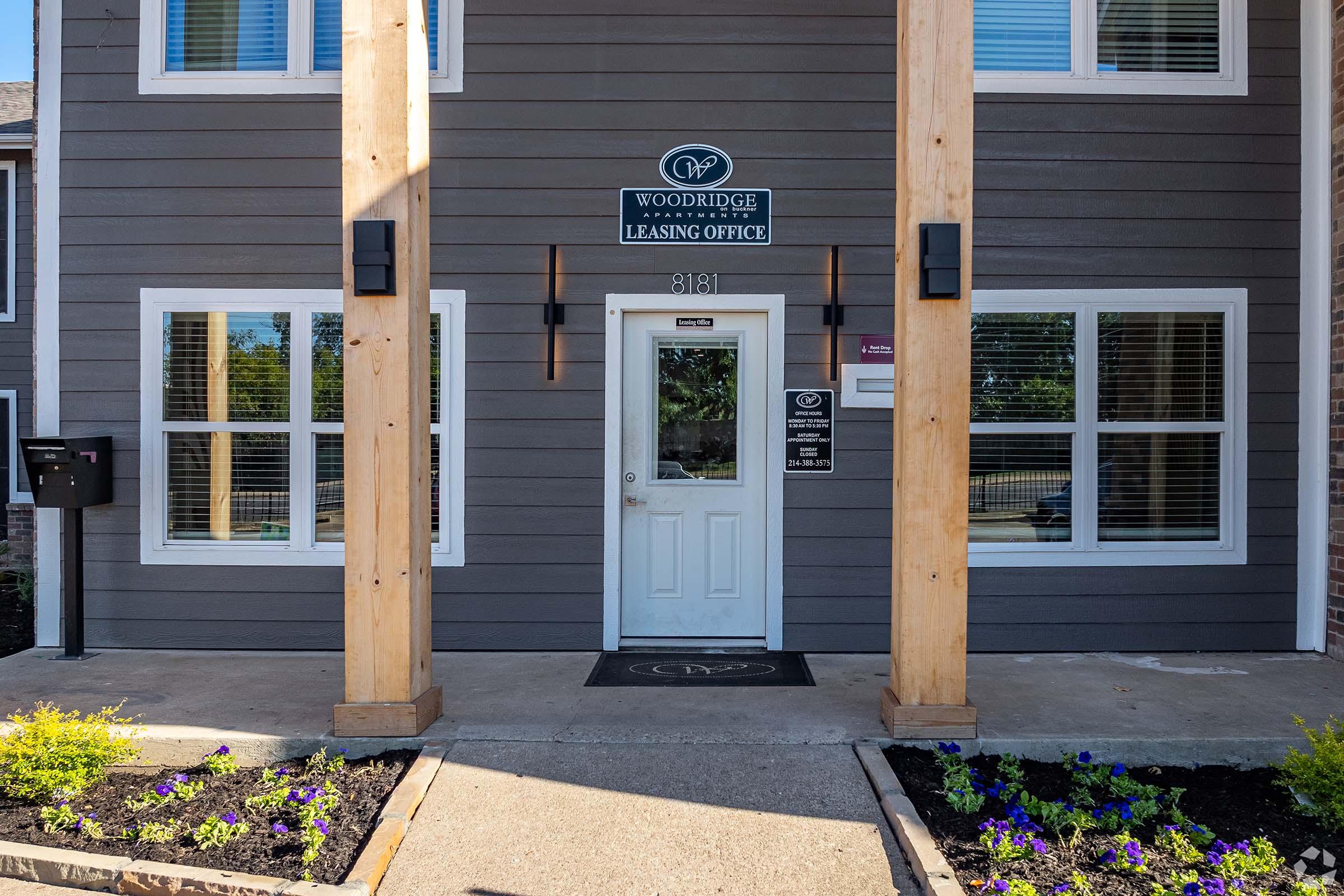 Entrance of the Woodridge Apartments Leasing Office, featuring a white door with a sign above, wooden support beams, and landscaped flower beds with small purple flowers. Mailbox is visible on the left. The building has a modern exterior with large windows.