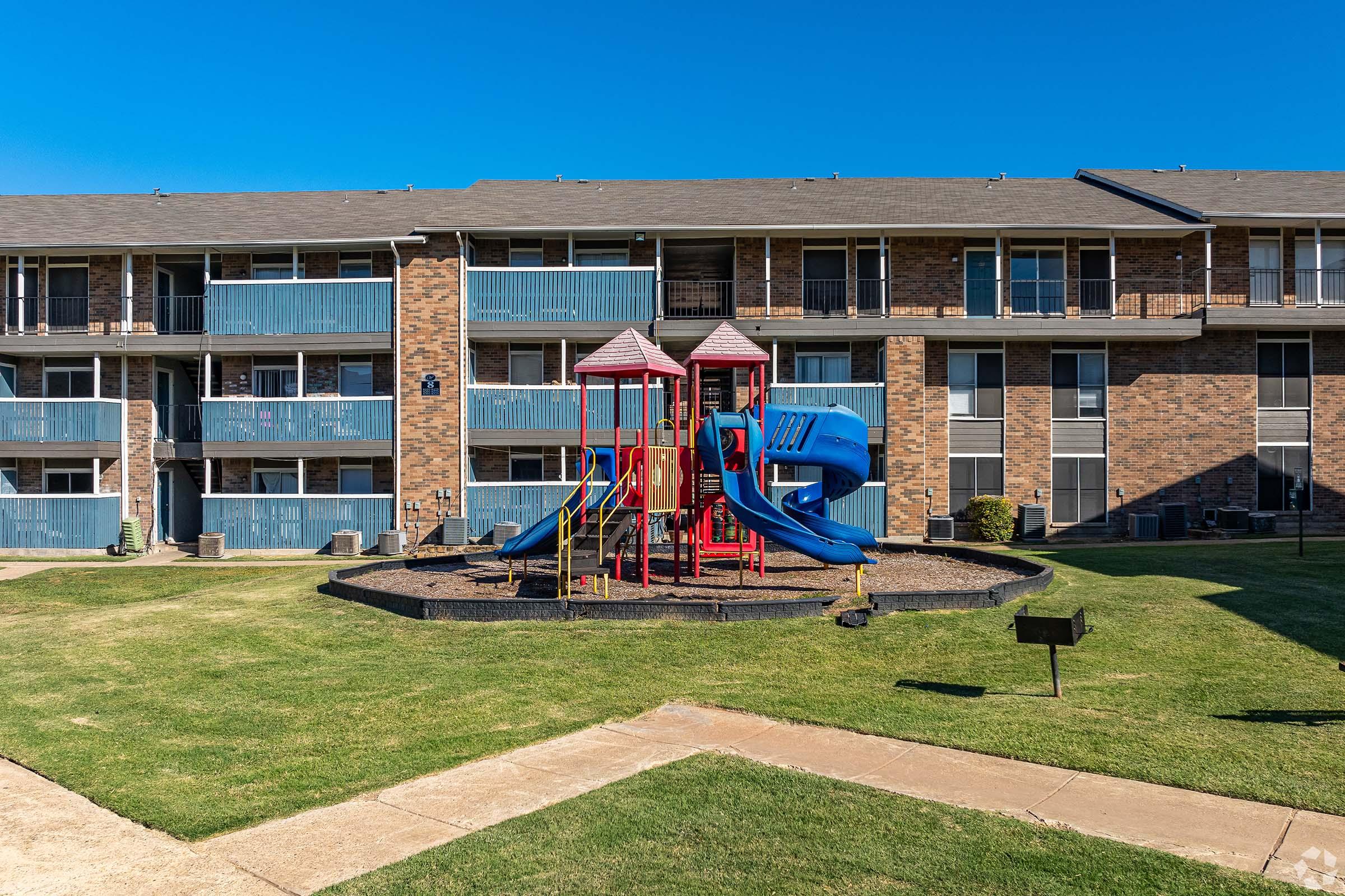 A colorful playground with slides and climbing structures sits on a grassy area in front of a multi-story brick apartment building. The clear blue sky and well-maintained surroundings create a cheerful setting for children to play.