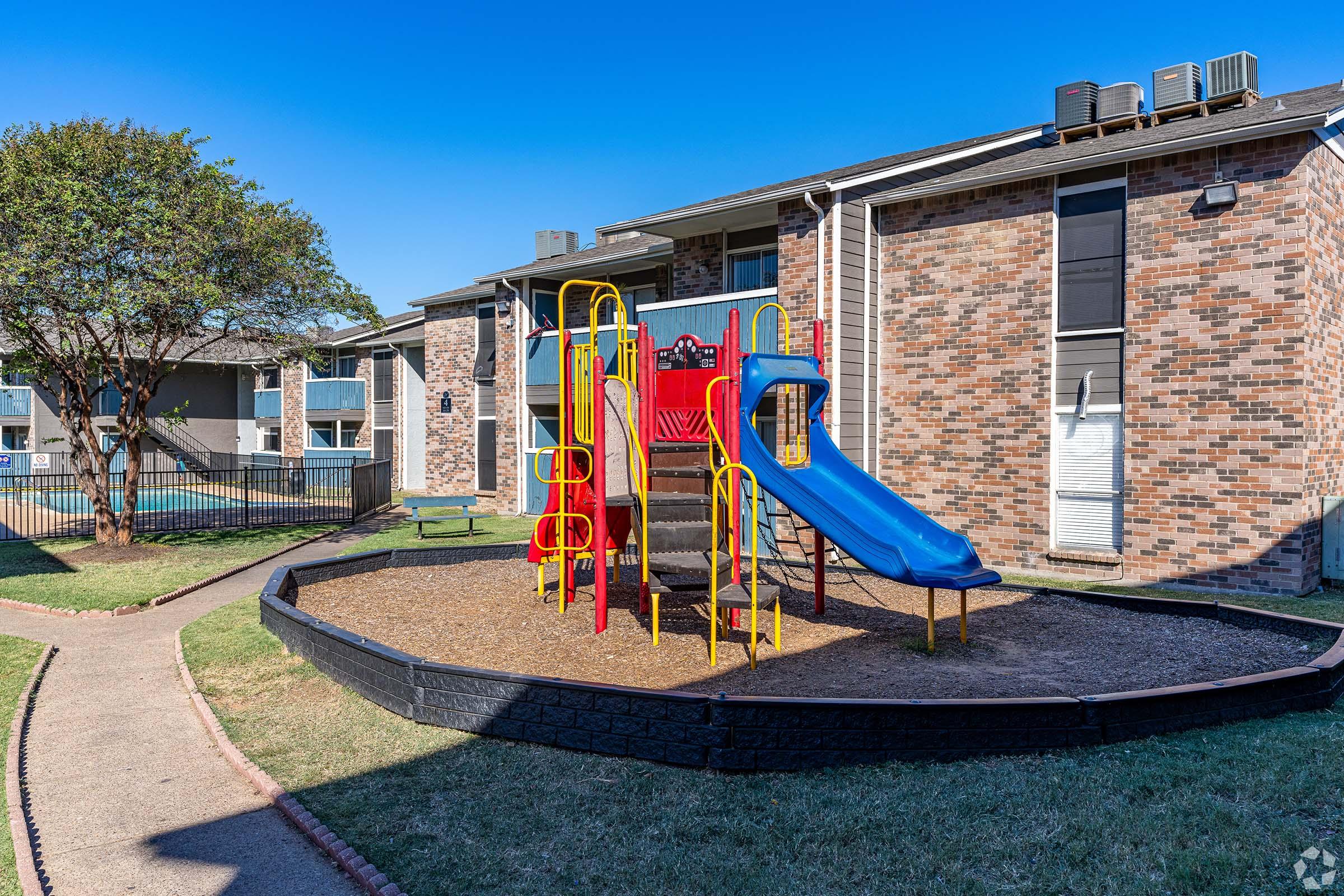 A colorful playground featuring a blue slide and climbing structures, surrounded by grassy areas and brick buildings. The playground includes safety logs and is situated in a well-maintained courtyard. Clear blue skies enhance the cheerful atmosphere.