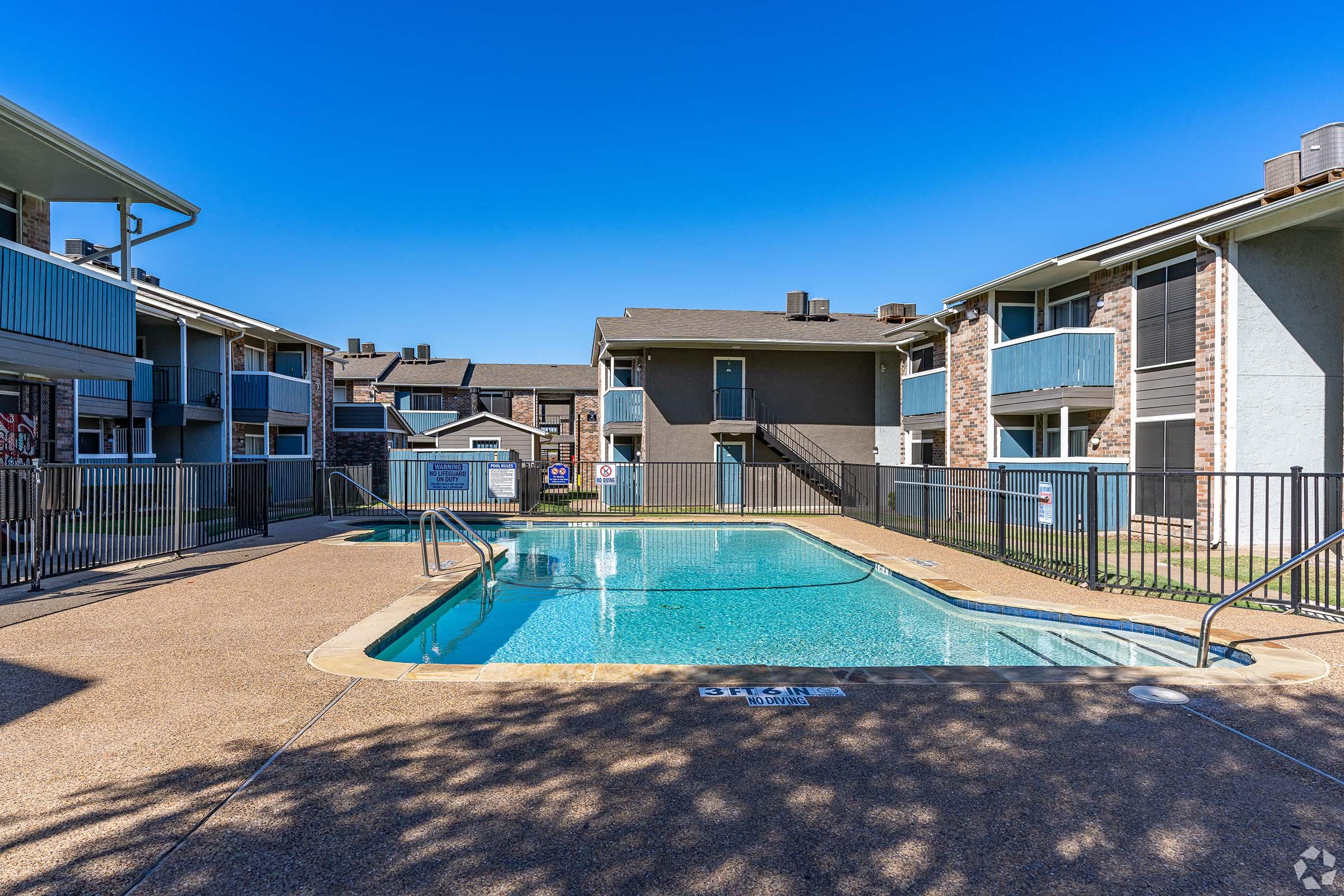 Swimming pool surrounded by a fence with two-story apartment buildings in the background. The pool deck is paved, and the water is clear and inviting. The sky is a bright blue, indicating a sunny day.