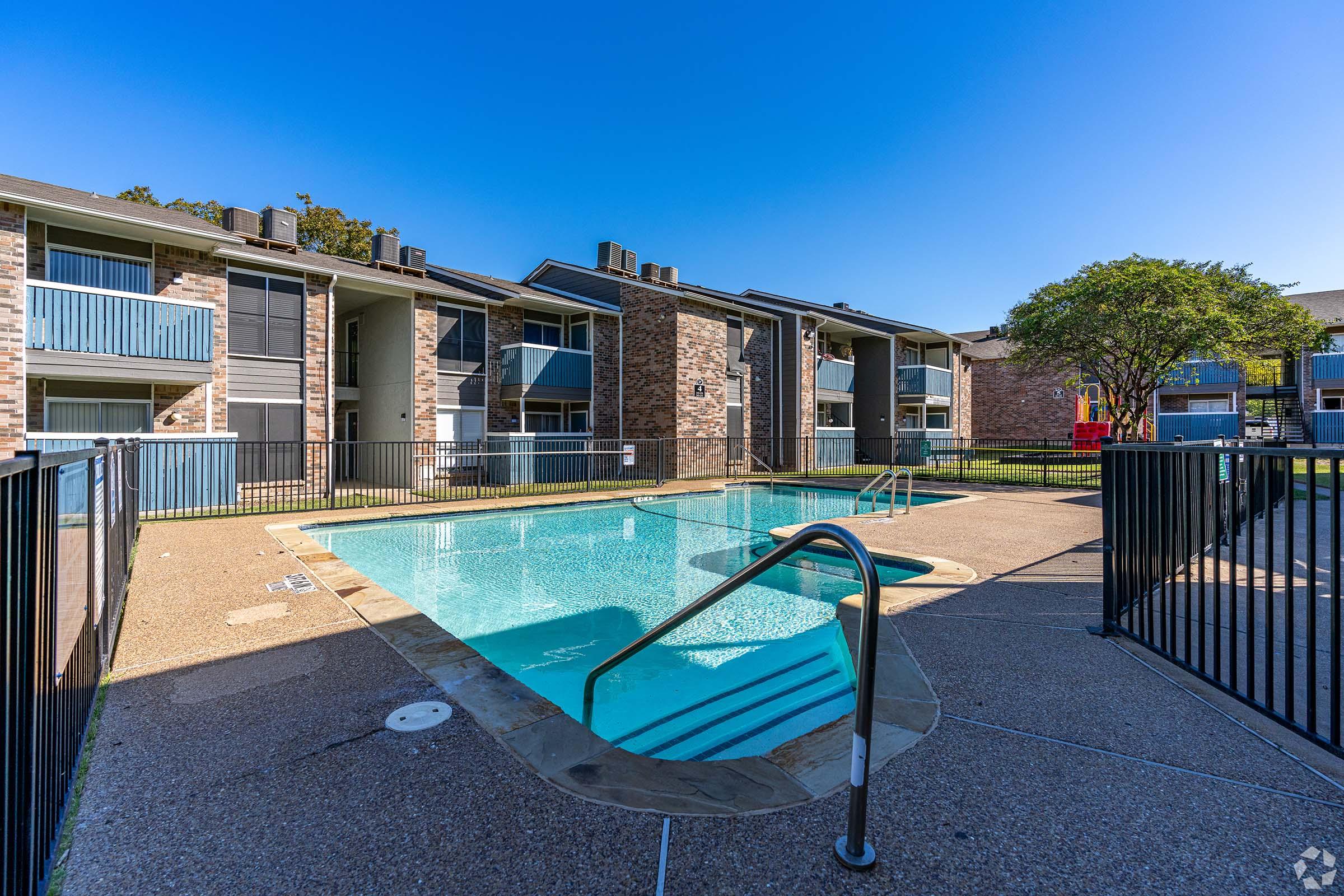 A well-maintained outdoor swimming pool surrounded by a fenced area, with two multi-unit apartment buildings in the background. The sky is clear and blue, and there are trees nearby, creating a pleasant residential atmosphere.