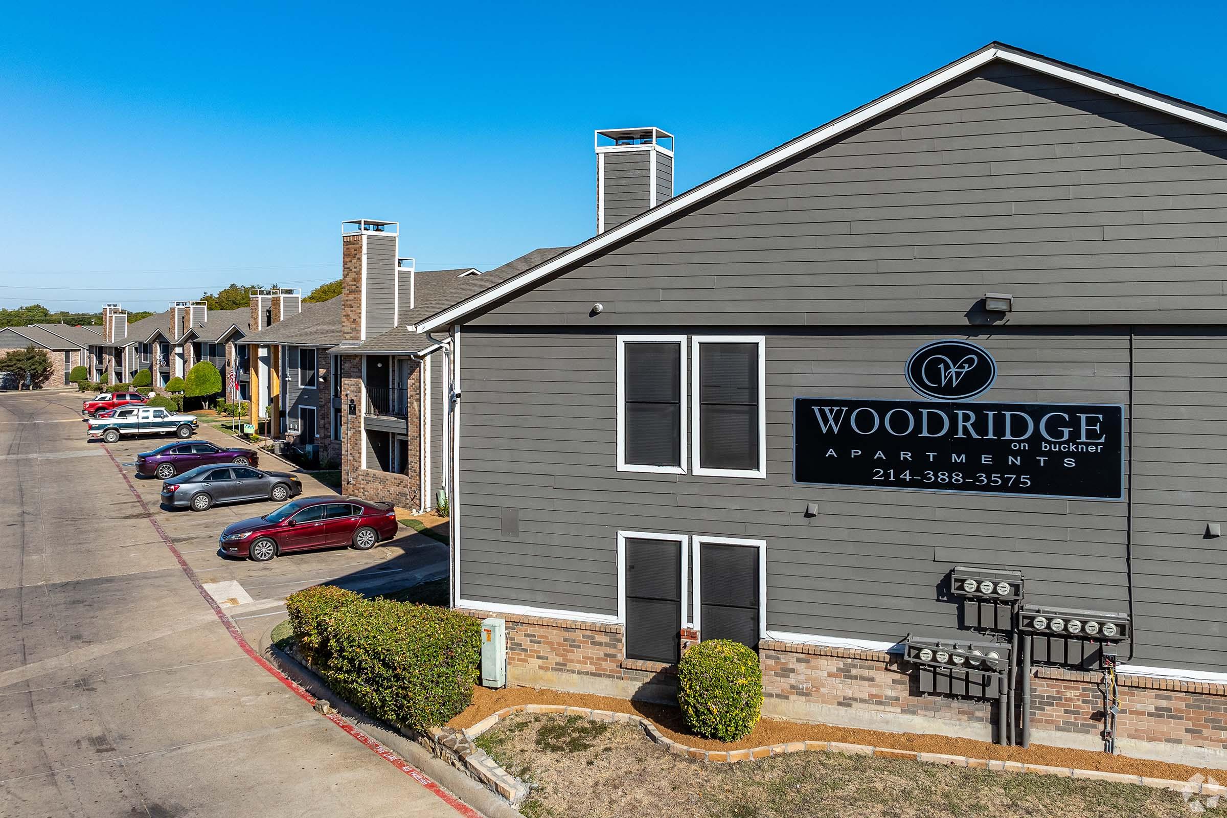 Exterior view of Woodridge Apartments on Butler, featuring multiple residential buildings. The image shows a well-maintained parking lot with several parked cars and landscaping. The sign with the apartment's name and contact number is prominently displayed on a gray building.