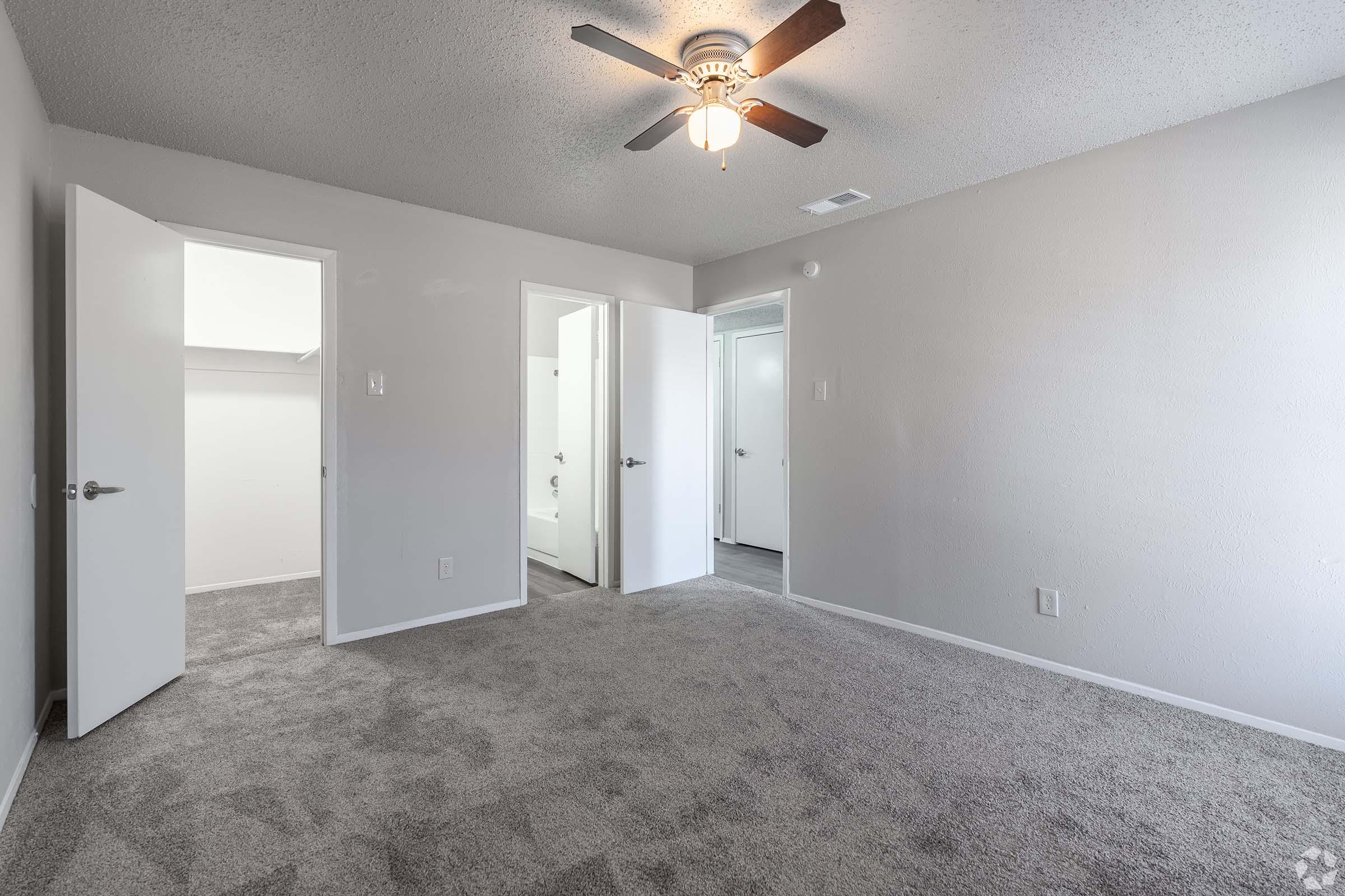 A well-lit, empty room with a light ceiling fan. The walls are painted in a neutral color, and there is soft gray carpet on the floor. Multiple doors lead to other spaces, including a bathroom. The overall ambiance is clean and modern, suitable for various uses.