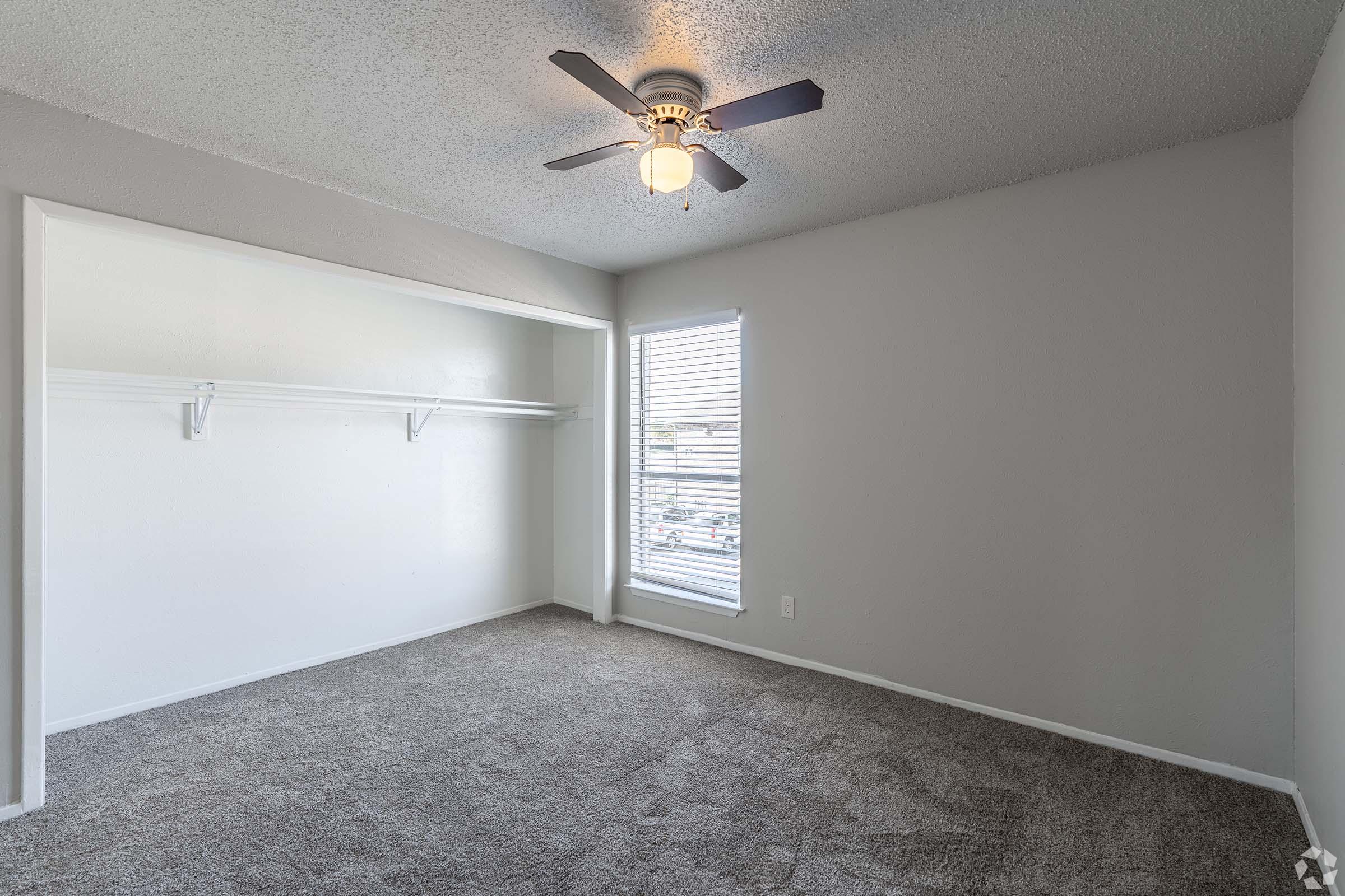 A spacious, empty bedroom featuring light gray walls and newly carpeted flooring. It has a ceiling fan, a small window with blinds, and a walk-in closet with shelving. Natural light filters in through the window, creating a bright and airy atmosphere.