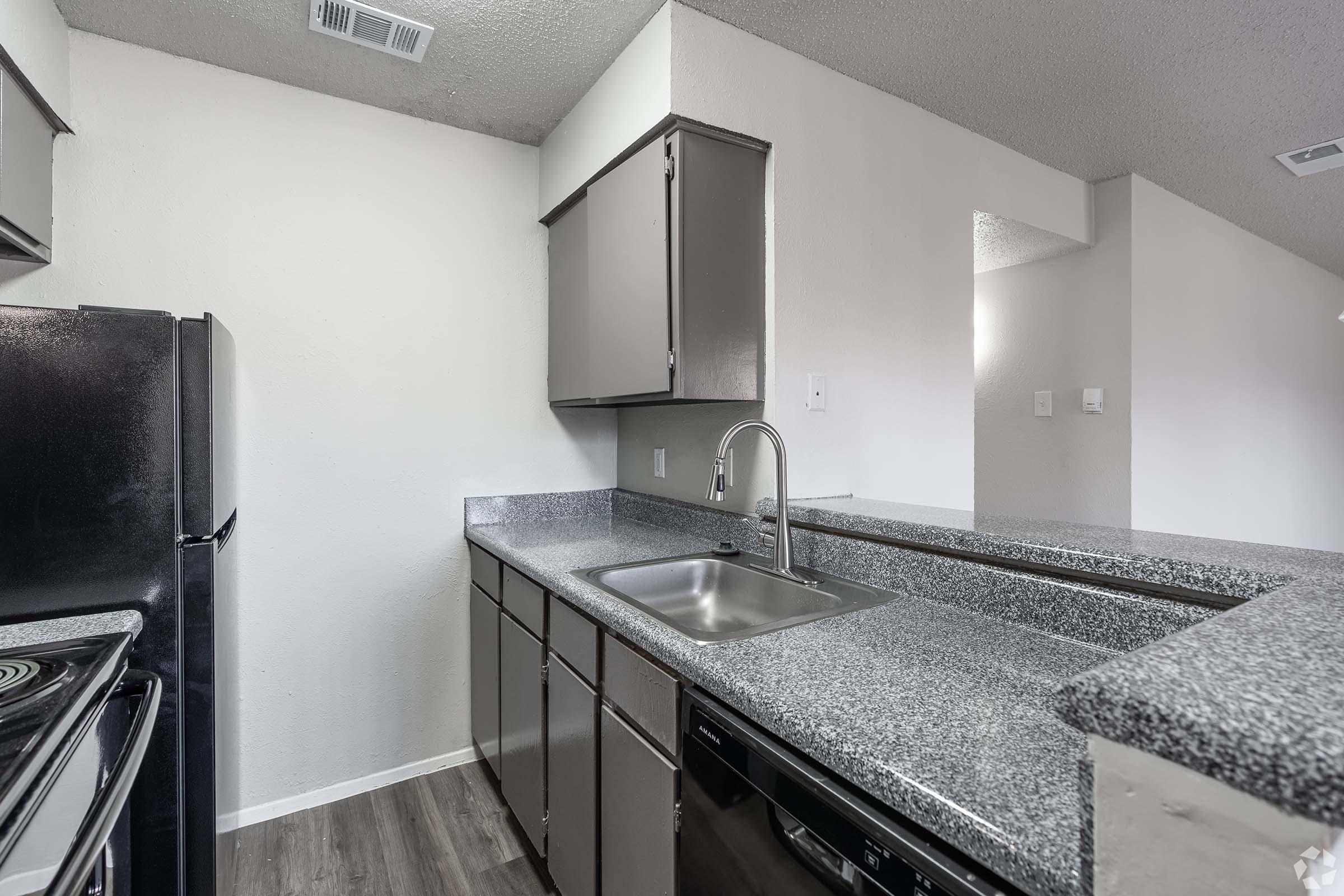 A modern kitchen featuring gray countertops, a stainless steel sink, and dark cabinets. It includes essential appliances like a black refrigerator and a built-in dishwasher. The design is open, with natural light visible from a nearby room, creating a spacious and inviting atmosphere.