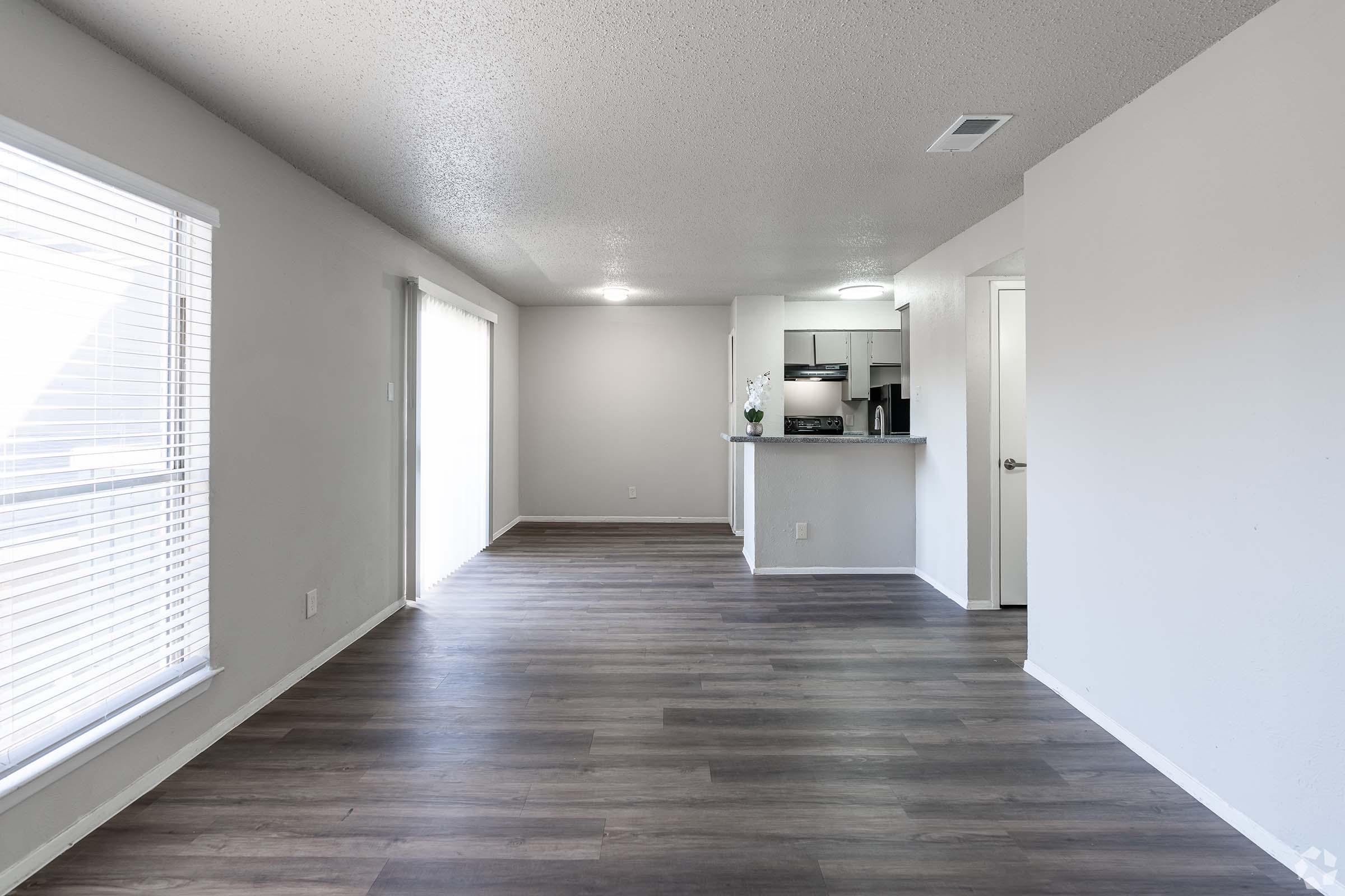 A spacious, well-lit interior showing an open living area with light-colored walls and modern vinyl flooring. To the left, a large window with blinds allows natural light, while the kitchen area is visible in the background. The overall ambiance is clean and contemporary.