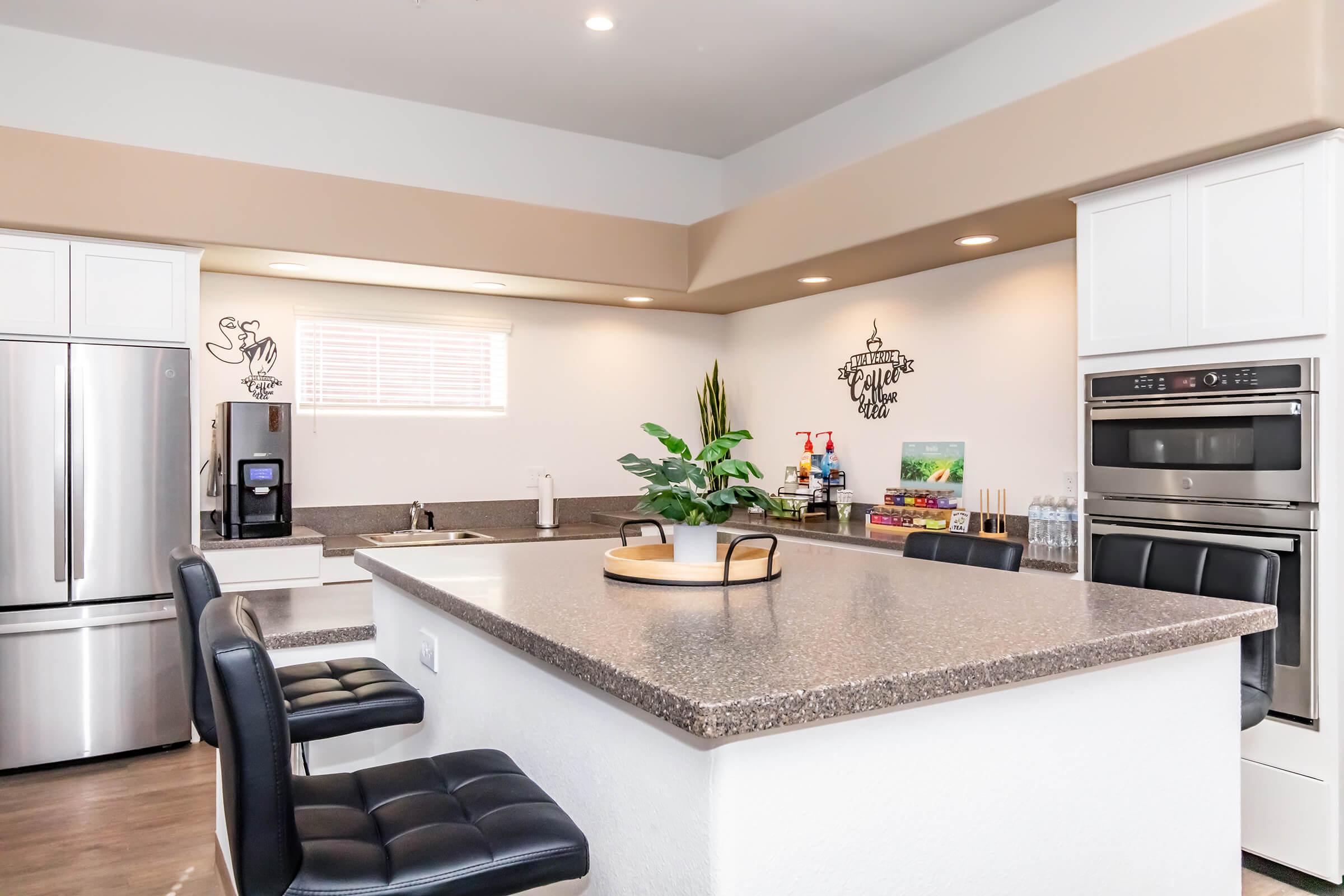A modern kitchen with a spacious island featuring a gray countertop. There are two black leather bar stools, stainless steel appliances including a refrigerator and an oven, and a window with natural light. Decorative items and plants are on the counter, adding a cozy touch to the space.