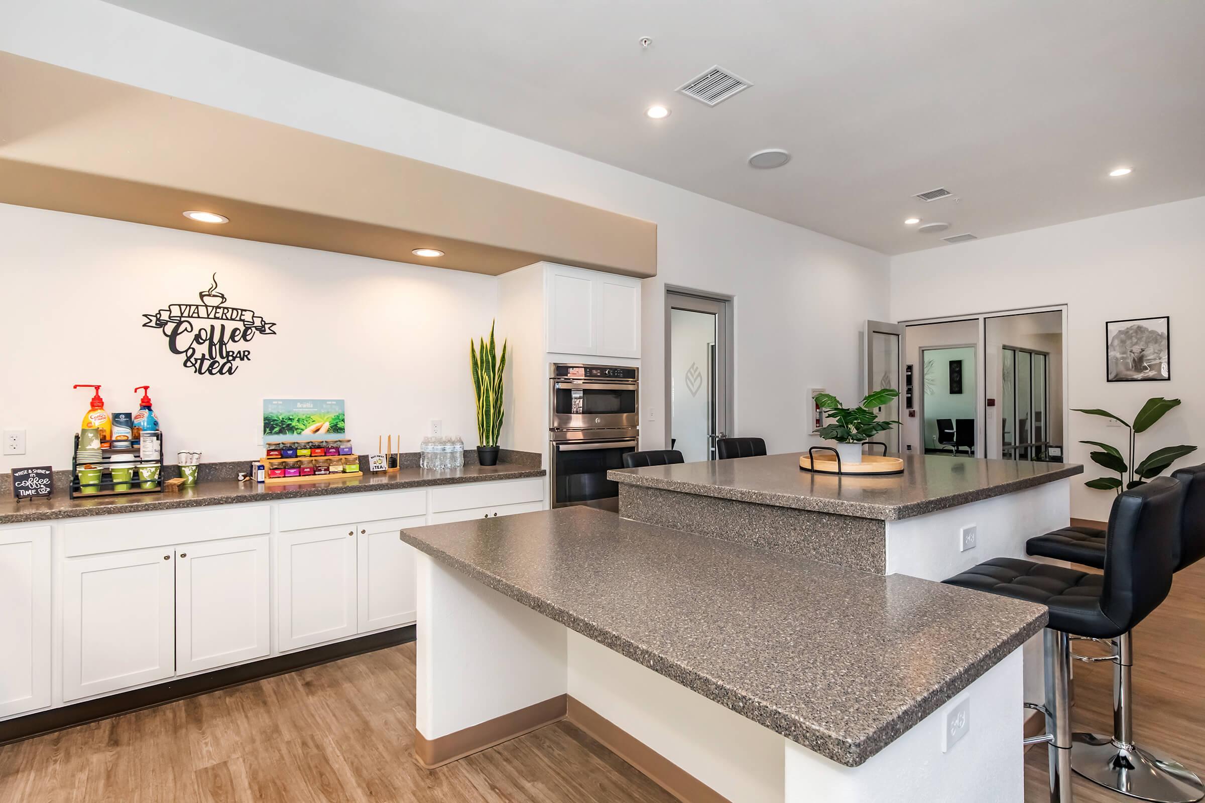 A modern kitchenette area featuring a large countertop with bar seating, coffee supplies, and decorative plants. The wall displays a decorative sign that reads "Welcome Home," creating a warm and inviting atmosphere. The open layout includes access to other rooms in a well-lit, contemporary space.