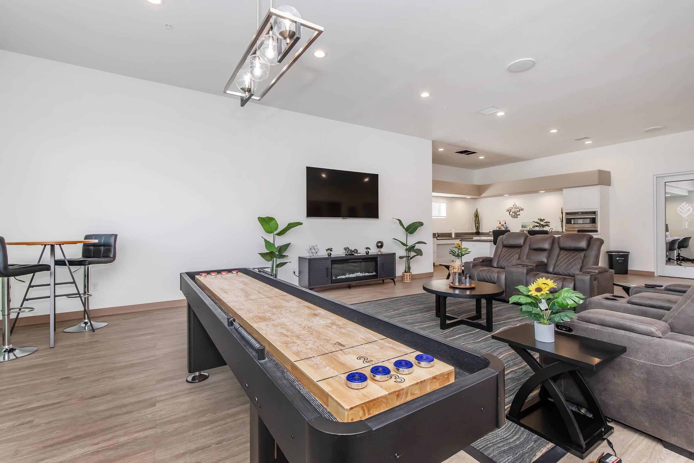 A modern living space featuring a shuffleboard table, a large flat-screen TV mounted on the wall, a cozy seating area with recliners, and stylish decor with plants. There is also a small bar with stools in the corner, enhancing the entertainment atmosphere of the room.