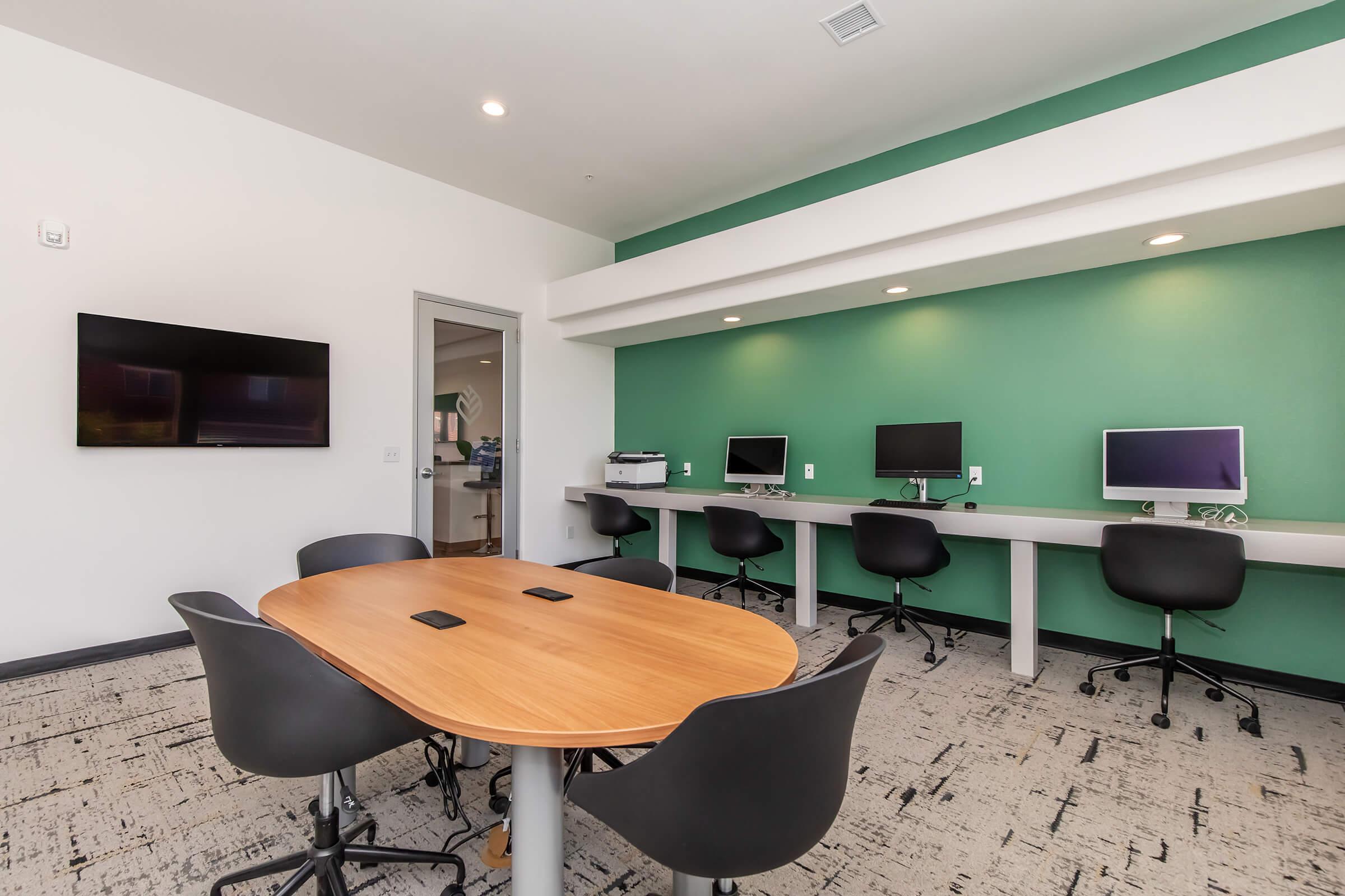 A modern conference room featuring a round wooden table surrounded by black chairs. Against one wall, there are two computers on white desks. A flat-screen TV is mounted on the wall, and the room is painted with a green accent wall, enhancing a professional and inviting atmosphere.