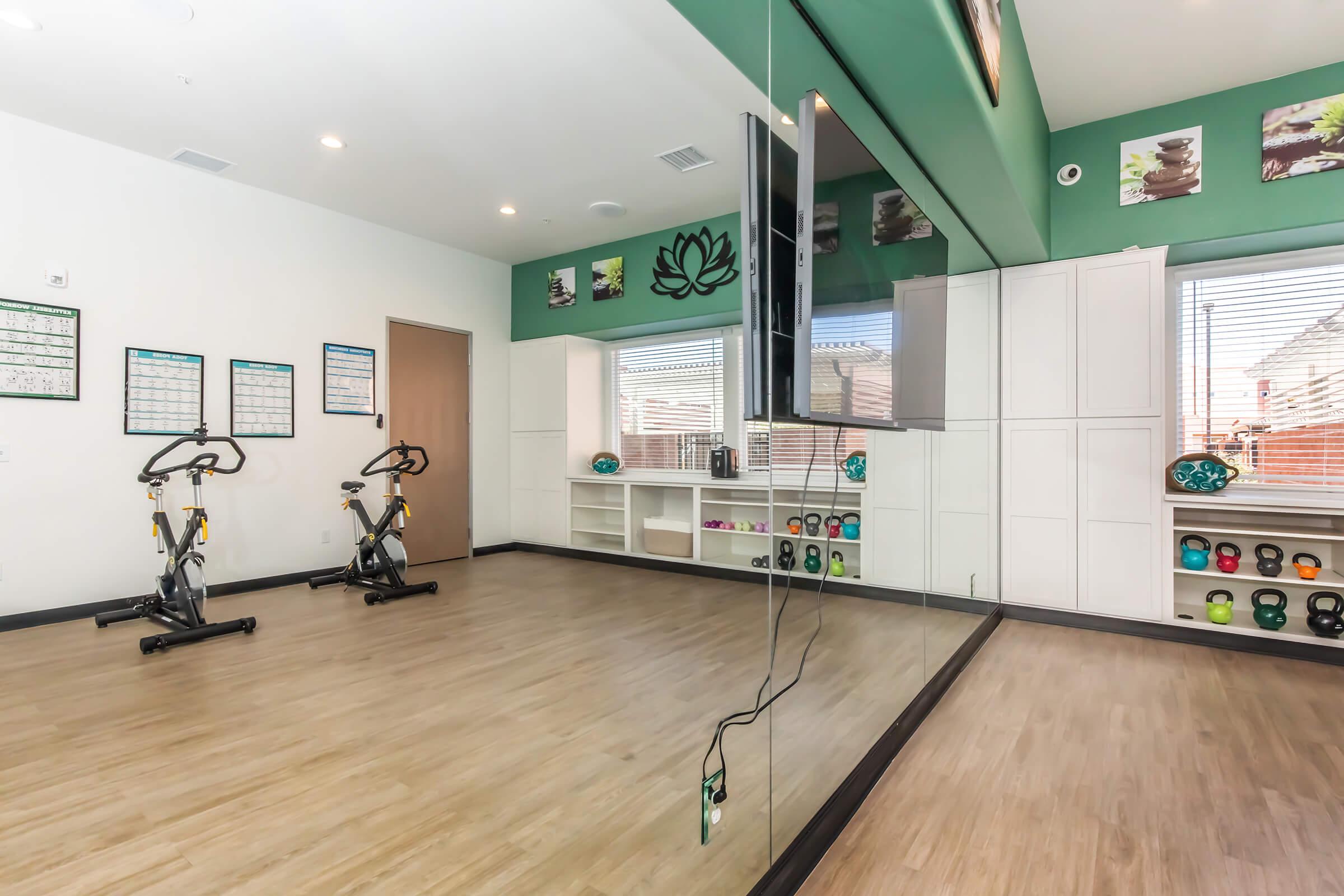 A well-lit fitness studio featuring two stationary bicycles, a large wall mirror, and shelves with colorful kettlebells. The walls are decorated with calming artwork, and there are calendars and motivational quotes displayed. Natural light enters through windows, enhancing the inviting atmosphere.