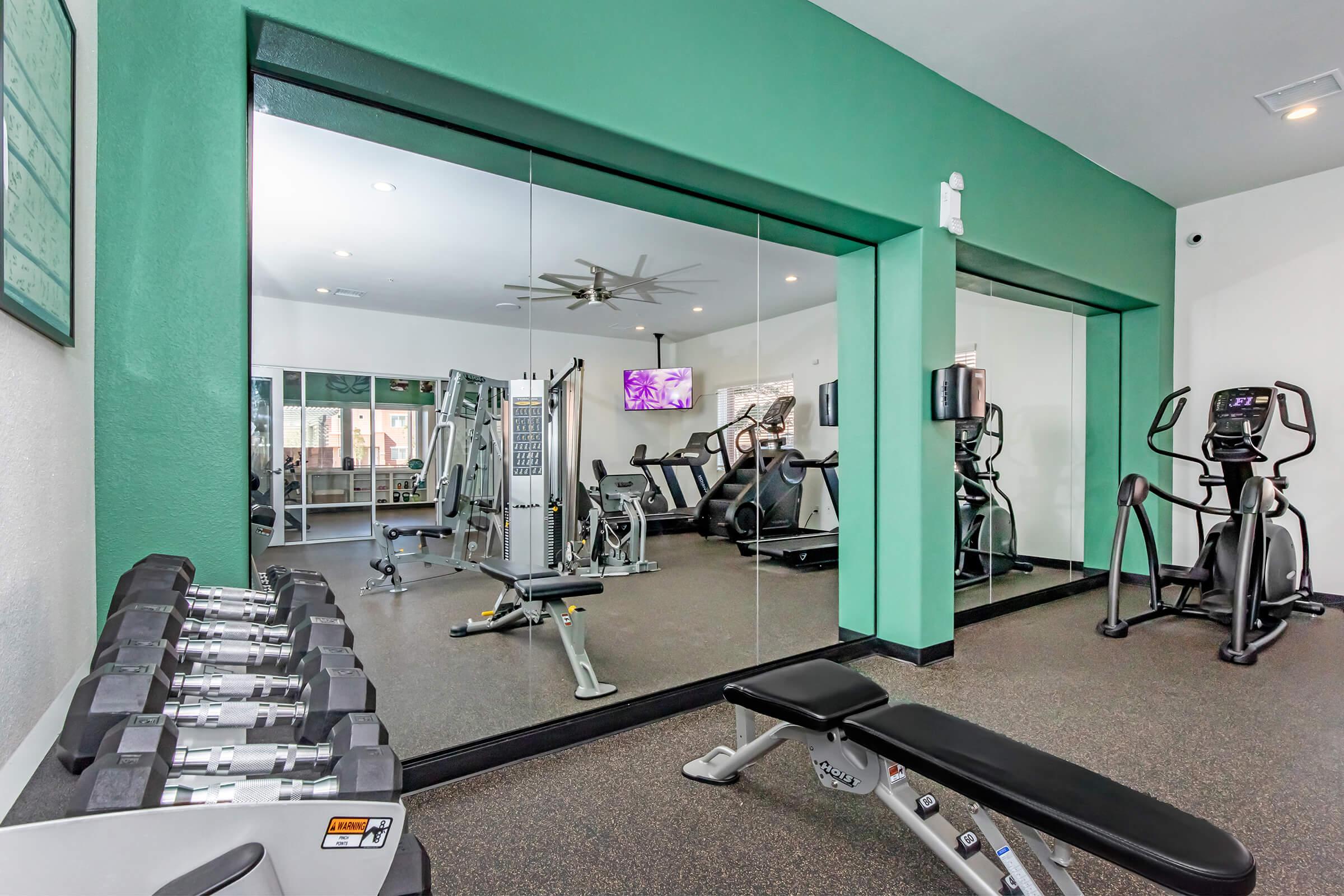 A fitness room featuring workout equipment including weights, an elliptical machine, and a bench, with large mirrors and natural light from windows. The walls are painted green, and there's a ceiling fan for ventilation. A television displays fitness content in the background.