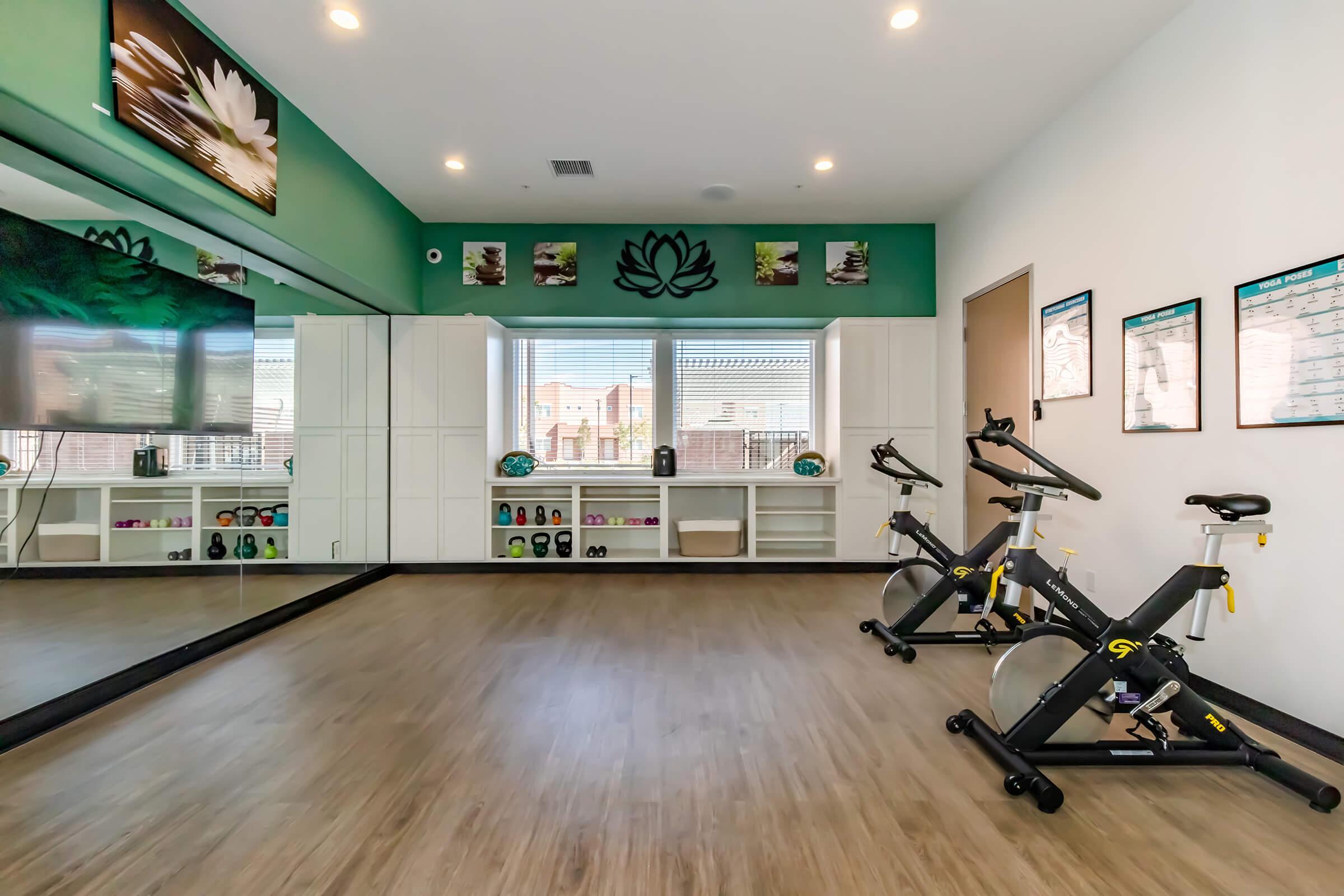 A bright and modern fitness studio featuring two stationary bikes, a large mirror on one wall, and a television. The space has green accent walls adorned with lotus artwork, and storage cabinets below windows that let in natural light. Fitness equipment and motivational posters are visible on the walls.