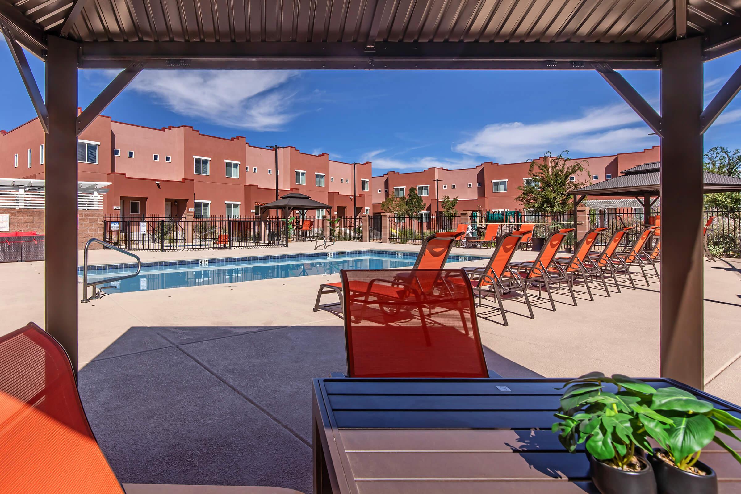 A view of a sparkling swimming pool surrounded by lounge chairs, with vibrant orange cushions. In the background, modern buildings are framed by clear blue skies and fluffy clouds. A small table with a potted plant is in the foreground, under a shaded structure.